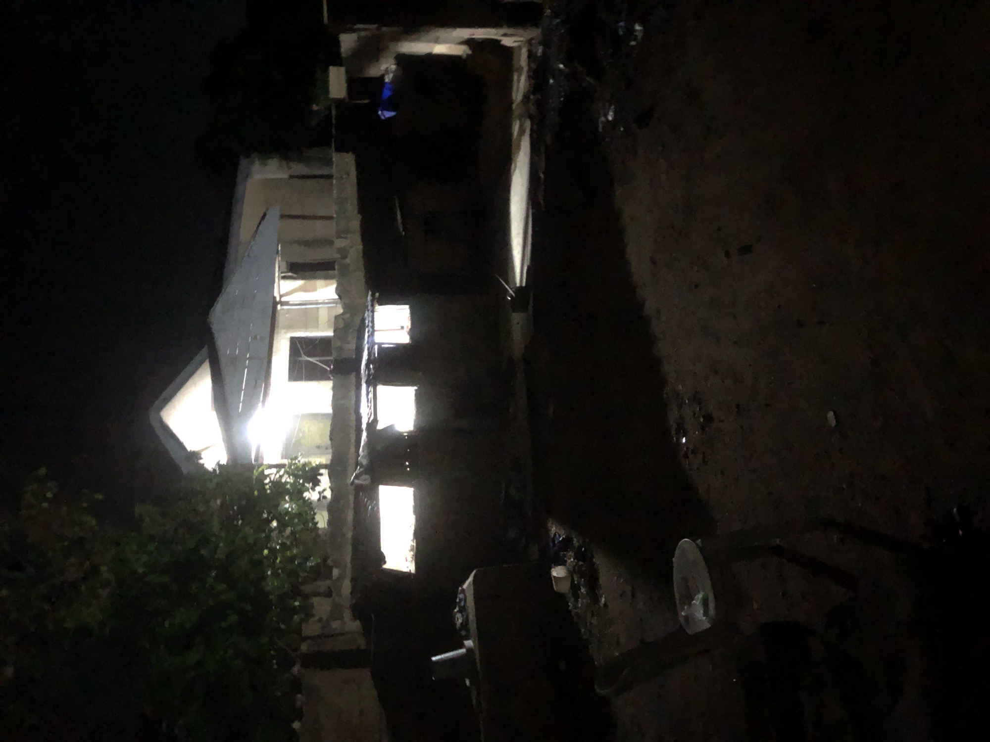 The image depicts an outdoor scene at night, illuminated by artificial lights. In the background, a building with a light-colored roof and walls is visible. Several windows in the building are brightly lit, suggesting interior lights are on. A large tree with dark foliage is situated to the left of the building. In the foreground and middle ground, there are several low-lying structures or walls, some of which have open, illuminated windows. A plastic chair with a light-colored seat is present in the lower left quadrant of the image. The ground appears to be rough and uneven, with scattered debris. The overall impression is a residential or semi-developed area at night in Kabong, Nigeria, with some structures appearing to be in a state of disrepair or under construction.
