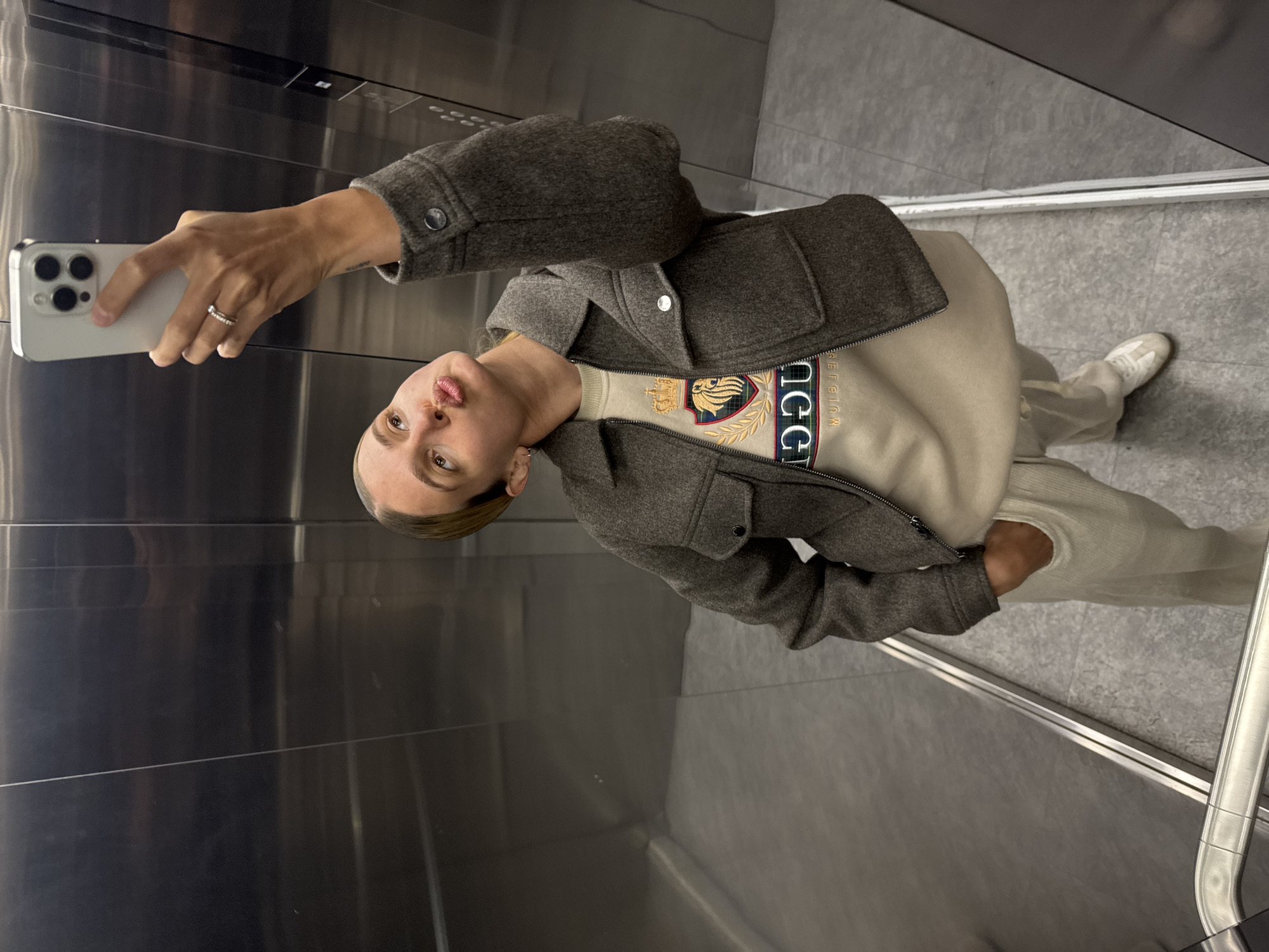 A young person takes a selfie in an elevator. The person wears a grey-brown jacket over a cream-colored sweater with a crest and light-colored trousers and shoes. They are holding a white smartphone with a visible triple-lens camera. The elevator interior is made of stainless steel with a grey floor.