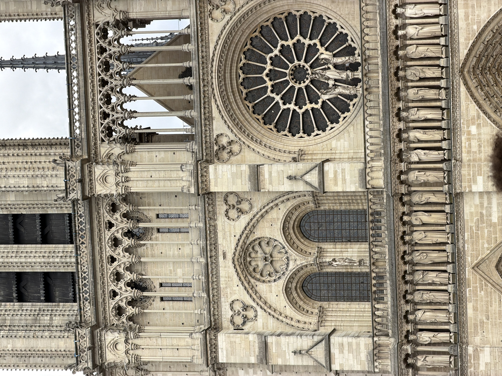 The image shows a close-up, vertical view of the facade of Notre Dame Cathedral in Paris, France. The architecture is primarily stone, with intricate carvings and details characteristic of Gothic style. Prominently featured is a large, circular rose window with delicate tracery and dark glass. Below the rose window are two arched windows with gridded panes. Along the right side of the image, a series of stone statues of figures in robes stand in niches, arranged in vertical rows. These statues and the surrounding architectural elements, such as columns, arches, and decorative friezes, are made of light-colored stone and exhibit significant detailing. To the left, parts of a spire and decorative elements of the upper facade are visible against a cloudy sky. The overall impression is of the detailed and weathered beauty of a historic religious structure. There is no visible text in the image. The lighting suggests it is daytime, but the cloudy sky indicates overcast weather.