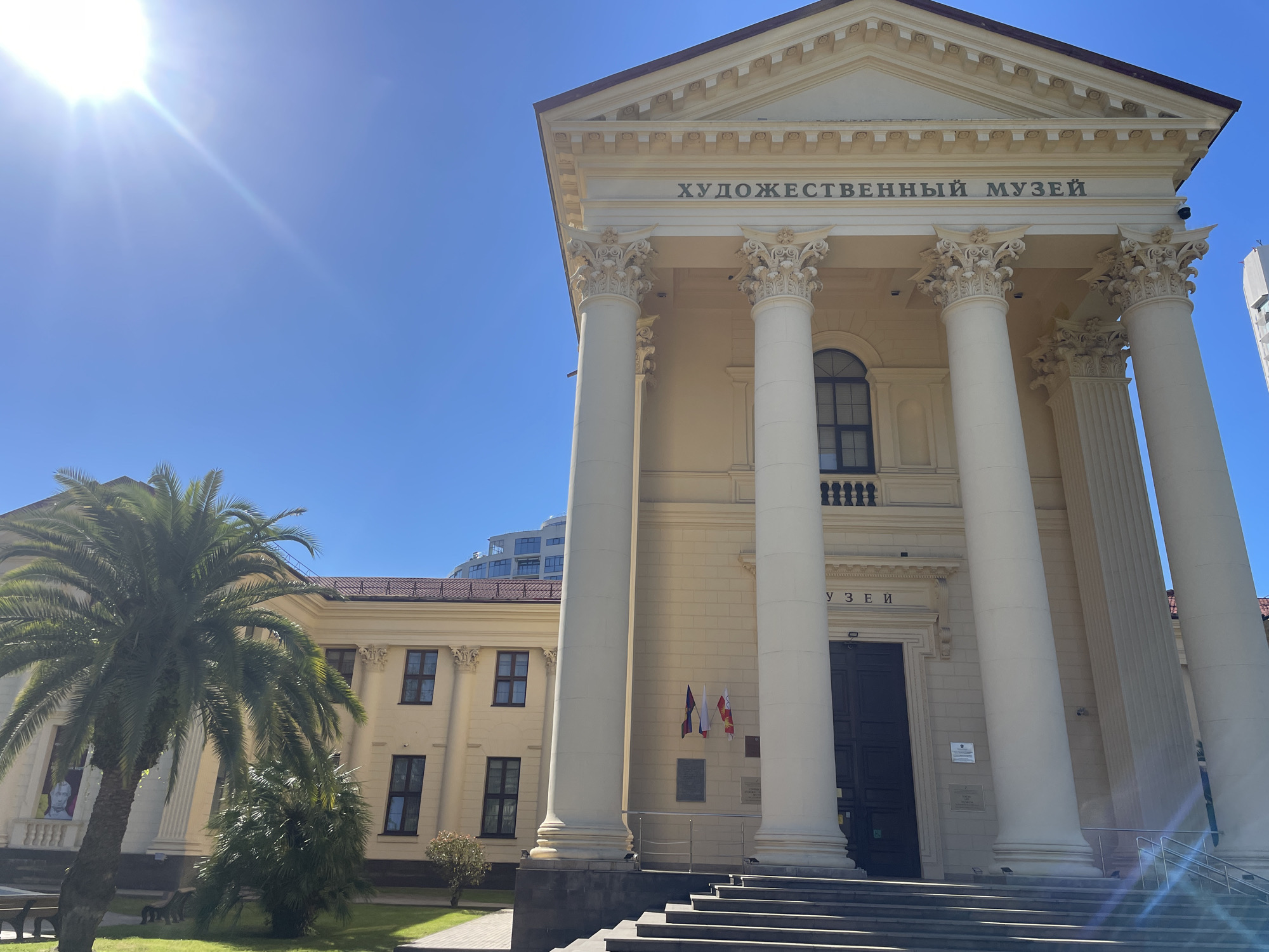 The image depicts the facade of a museum in Sochi, Russia, identified by the Russian text "ХУДОЖЕСТВЕННЫЙ МУЗЕЙ" (Art Museum) on its pediment. The architecture is classical, characterized by large white columns supporting a triangular pediment. In front of the museum, a grand staircase leads up to the entrance. To the left of the main building, a separate, lower structure with several windows is visible. A large palm tree stands prominently in the foreground on the left, with a smaller, bush-like tree to its right. The scene is bathed in bright sunlight, casting distinct shadows and creating lens flare from the sun, which is positioned in the upper left corner of the frame. The sky is clear and blue. No people are visible in the image.