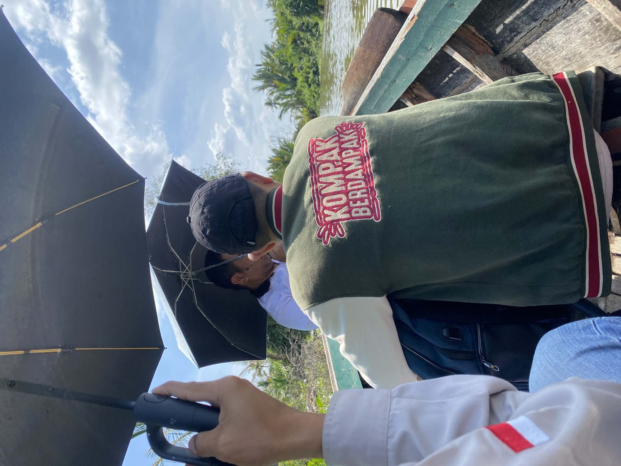 A group of people are in a boat on a river under a cloudy sky. A person in the foreground is holding an umbrella. Another person in the middle ground is wearing a green jacket with "KOMPAK BERDAMPAK" embroidered on the back. Lush vegetation lines the riverbanks, suggesting a rural or semi-rural setting. The boat appears to be made of wood, with visible wear and tear. The context of the image suggests a trip or journey on the river, possibly in a tropical region. The phrase "KOMPAK BERDAMPAK" is Bahasa Indonesia and translates to "Solidary Impacts."