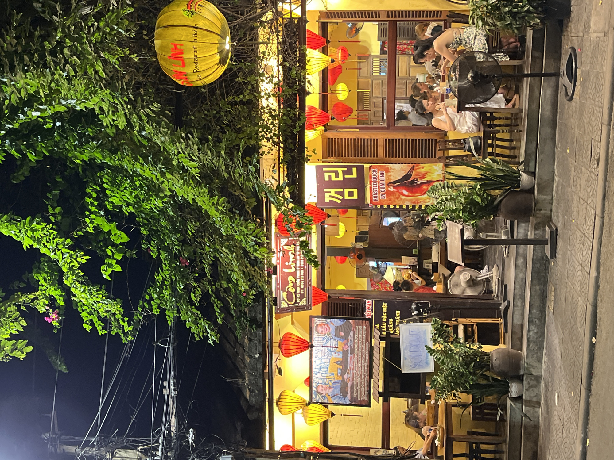 The image shows a restaurant in Hội An Ward, Vietnam at night. The restaurant exterior features yellow walls decorated with traditional hanging lanterns. Diners are seated both inside and outside the restaurant, with some tables visible on a raised platform extending onto the sidewalk. Fans provide cooling for the diners. Restaurant signage includes names like "Cơm Linh" and advertises "ROASTED DUCK BY COM LINH". A yellow spherical lantern hangs from a tree near the restaurant entrance. Power lines and foliage are visible in the foreground.