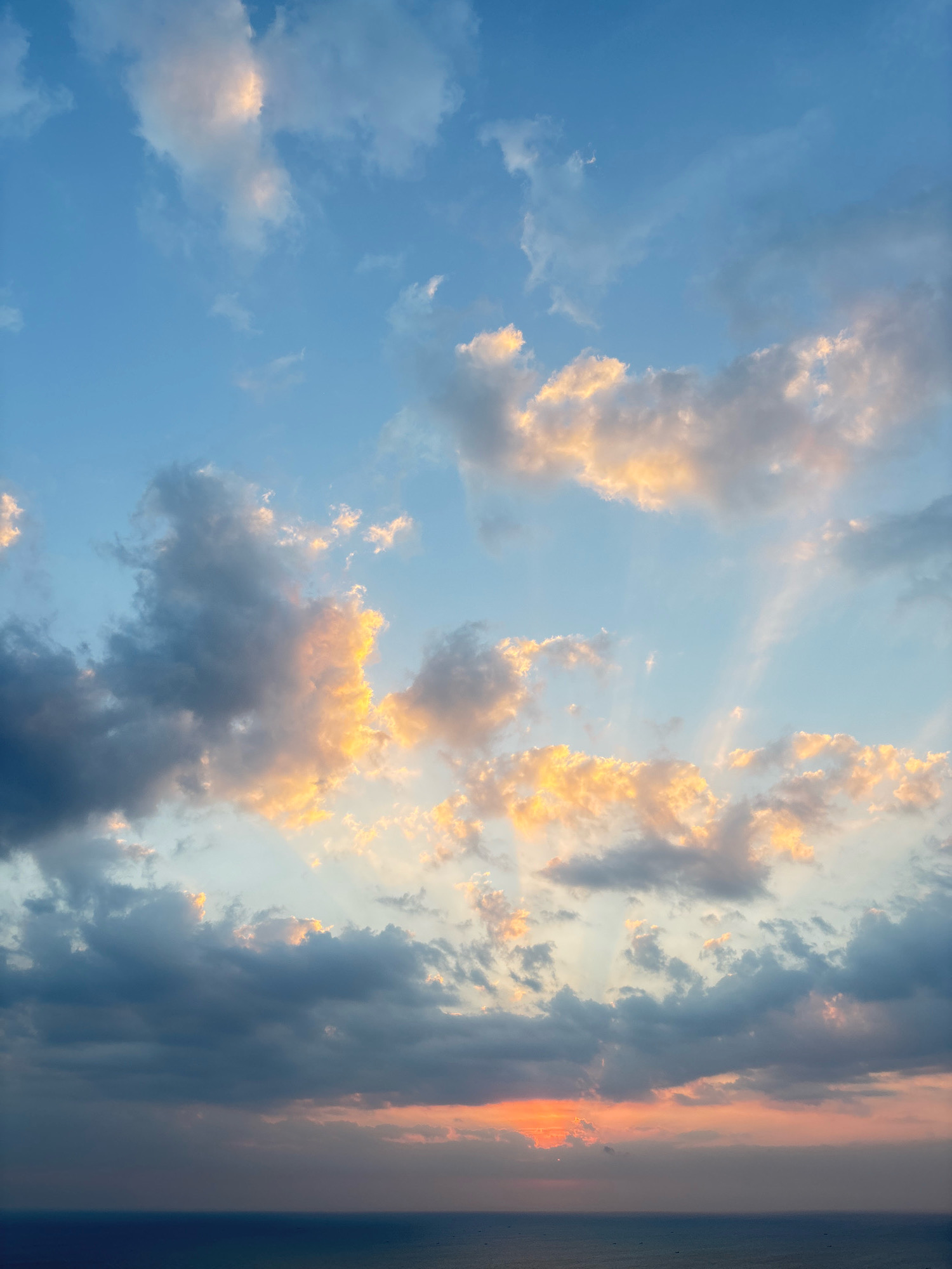 The image depicts a sunset over the ocean, as seen from Kuta Selatan, Indonesia. The sky is filled with a mixture of blue, gray, and orange clouds, with the sun a faint orange glow peeking through the clouds near the horizon. The ocean is a deep blue, stretching out to meet the sky. The scene is tranquil and serene, with no people or objects visible in the frame, creating a sense of vastness and natural beauty.