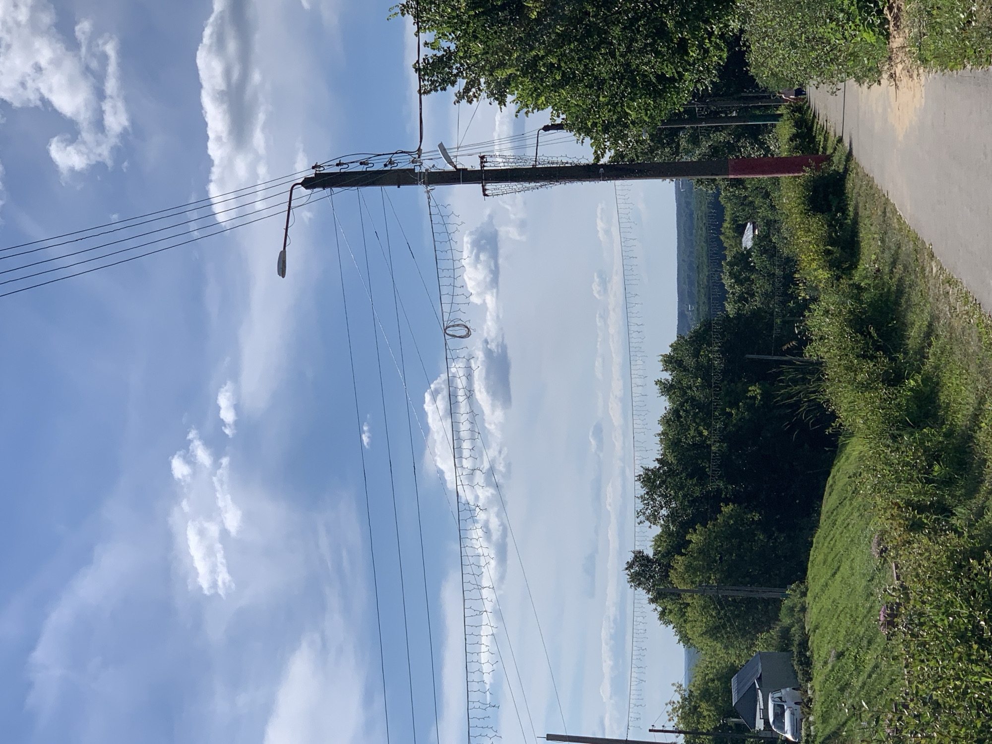 This is a medium-shot, outdoor photograph taken on a sunny day with scattered clouds in the sky. The scene appears to be a rural or suburban area on the outskirts of Pavlovo, Russia, likely during summer.
In the foreground, a paved pathway runs diagonally from the lower right to the mid-left of the frame. The pathway is bordered on the right by dense green bushes and trees. To the left of the pathway, a grassy slope rises and is covered with various plants. A white van is parked on the left side of the slope, near a small building with a dark roof.
Dominating the mid-ground and background is a dense expanse of green trees, indicating a wooded or heavily forested area. In the distance, the landscape appears to undulate, suggesting rolling hills or a valley.
Several utility poles with overhead wires are visible throughout the image, including one prominent pole in the mid-right with a street lamp attached and a red-painted section near its base. These poles support numerous electrical wires, with a string of decorative lights draped between some of them.
A lone figure, appearing to be a person walking, is visible on the pathway in the distance towards the right side of the image. The lighting suggests it is daytime, with the sun casting shadows. The overall impression is of a tranquil, natural setting with signs of human infrastructure.