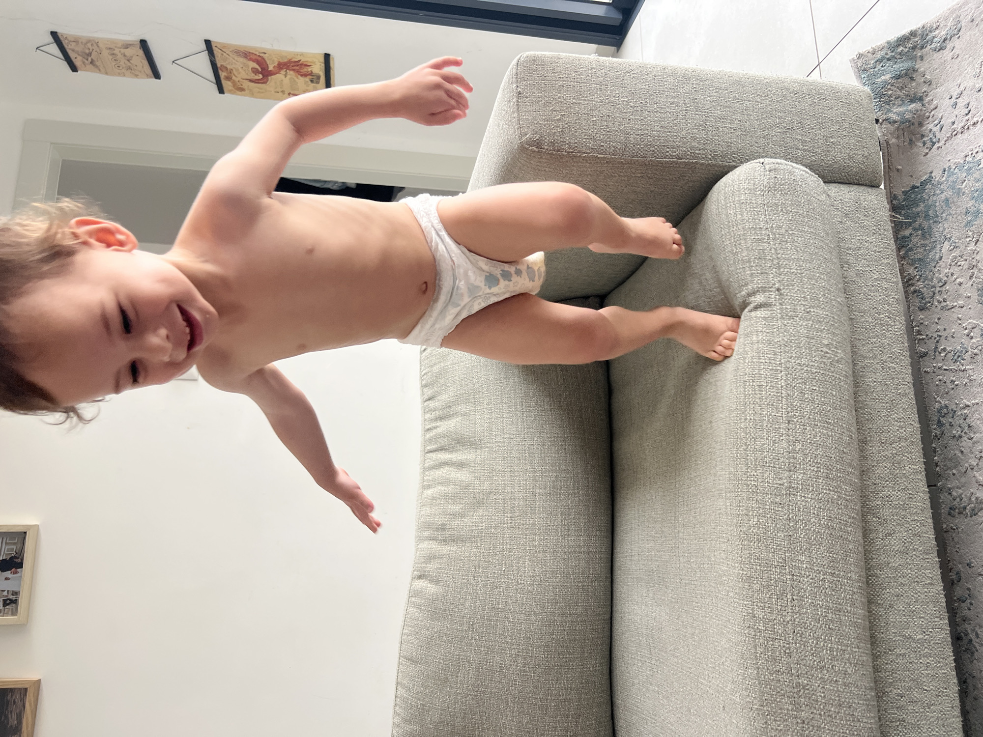 A toddler wearing a diaper is playing on a light gray sofa. The room has white walls, a white door frame, and hanging art. The scene is likely indoors. The location is Kiryat Bialik, Israel, based on information not included in the image.