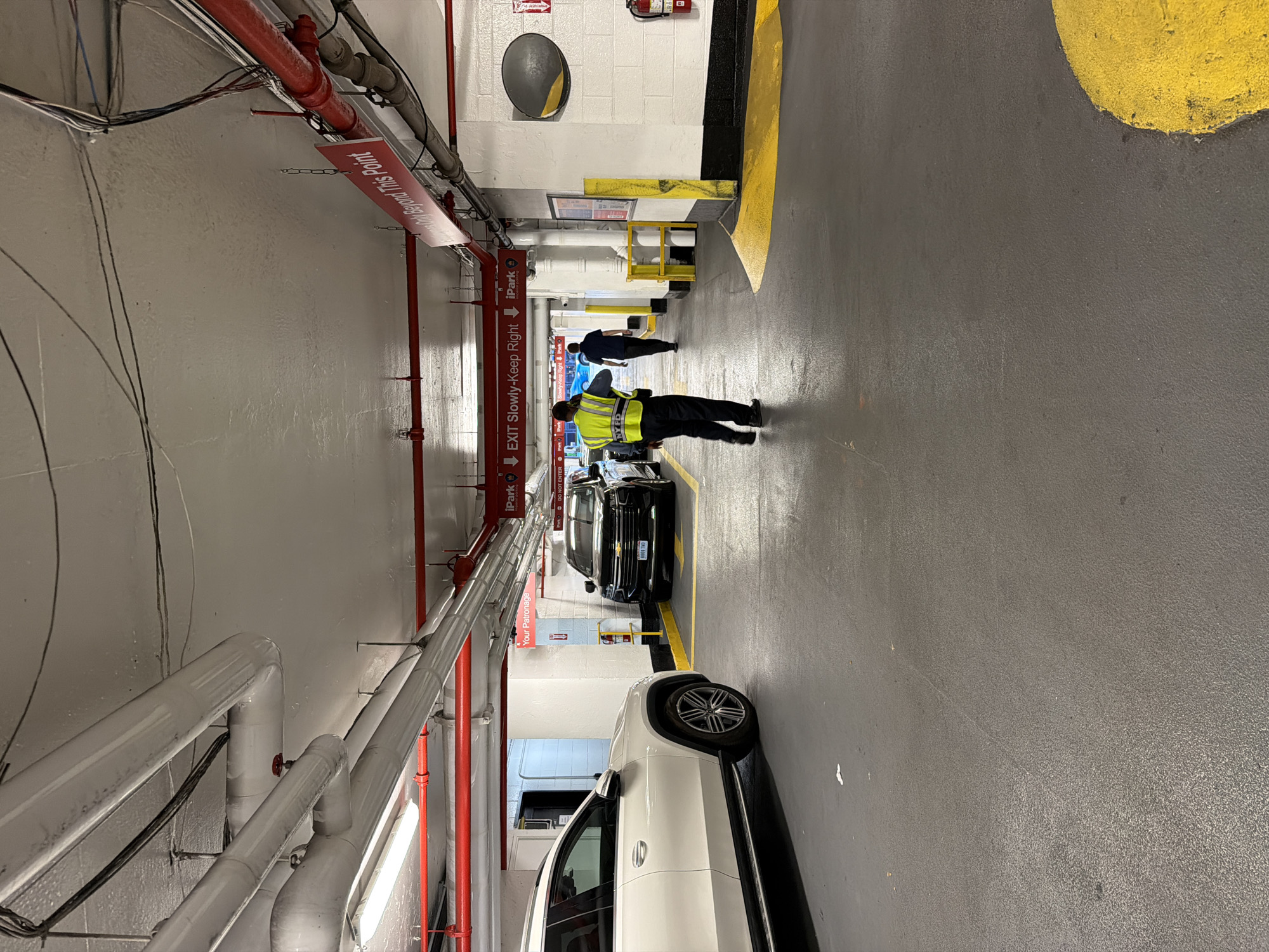 A security guard in a yellow vest and dark pants, identified by "NYFD" on his vest, walks away from the camera in a parking garage. He is in the process of exiting a parking spot and walking down a lane. Behind him, a dark colored SUV is parked in a spot marked by yellow lines. Further down the lane, another person in a dark shirt and pants walks away from the camera. The garage has a concrete ceiling with exposed pipes and fluorescent lighting. Red pipes are visible overhead, along with various wires. Signs are mounted on the ceiling, including one that reads "EXIT SLOWLY - KEEP RIGHT" and another that says "iPark". A white SUV is parked to the left of the frame. The floor of the garage is grey asphalt with yellow markings. A fire extinguisher is mounted on the wall to the right of the frame. The scene appears to be taking place in a parking garage located in New York City, United States of America.