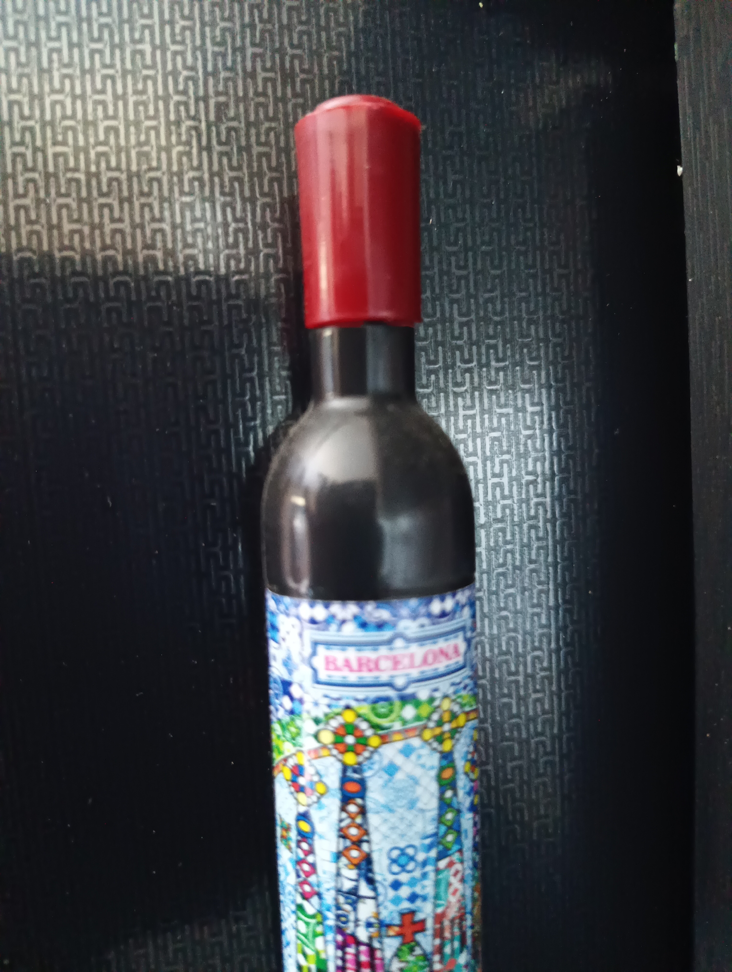 A vertical, eye-level shot shows a black bottle with a red cap. The bottle has a decorative label with the word "BARCELONA" in a light blue rectangle with a white border. The label features a mosaic pattern in various colors, including blue, white, red, yellow, and green, with geometric shapes and a stylized representation of architecture. The background is a dark, textured surface with a repeating pattern of interconnected geometric shapes, possibly a textured wall or fabric. The lighting casts a shadow on the left side of the bottle and the background. The location of Karnobat, Bulgaria is not depicted or suggested in this image.