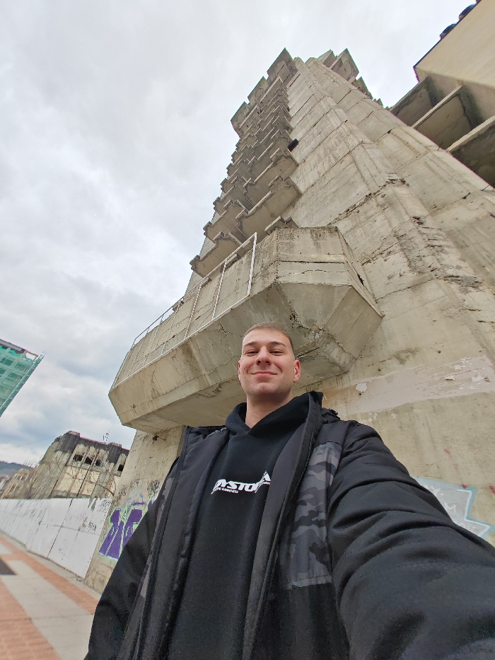 A male individual is depicted in a low-angle, wide-perspective selfie in Shumen, Bulgaria, positioned on a paved pedestrian surface. The subject is a young adult male with light skin, short-cropped hair, and a faint smile, looking directly at the camera. He wears an open black jacket with a camouflage pattern on the shoulders over a black hooded sweatshirt featuring white text, partially visible as "ASTOR" or "ASTORM" above "ESS". Behind and to the right of the subject, an imposing, multi-story, unfinished concrete high-rise building dominates the frame, extending vertically into an overcast sky. This structure, known as the "unfinished hotel" in Shumen, displays exposed, weathered concrete with staggered, projecting balconies or floor slabs and some corroded metal railings on its lower sections. To the left, a concrete wall with purple graffiti is visible. Further in the background, another building under construction is partially obscured by green scaffolding mesh, and other distant, dilapidated structures are visible under the uniformly gray, cloudy sky.