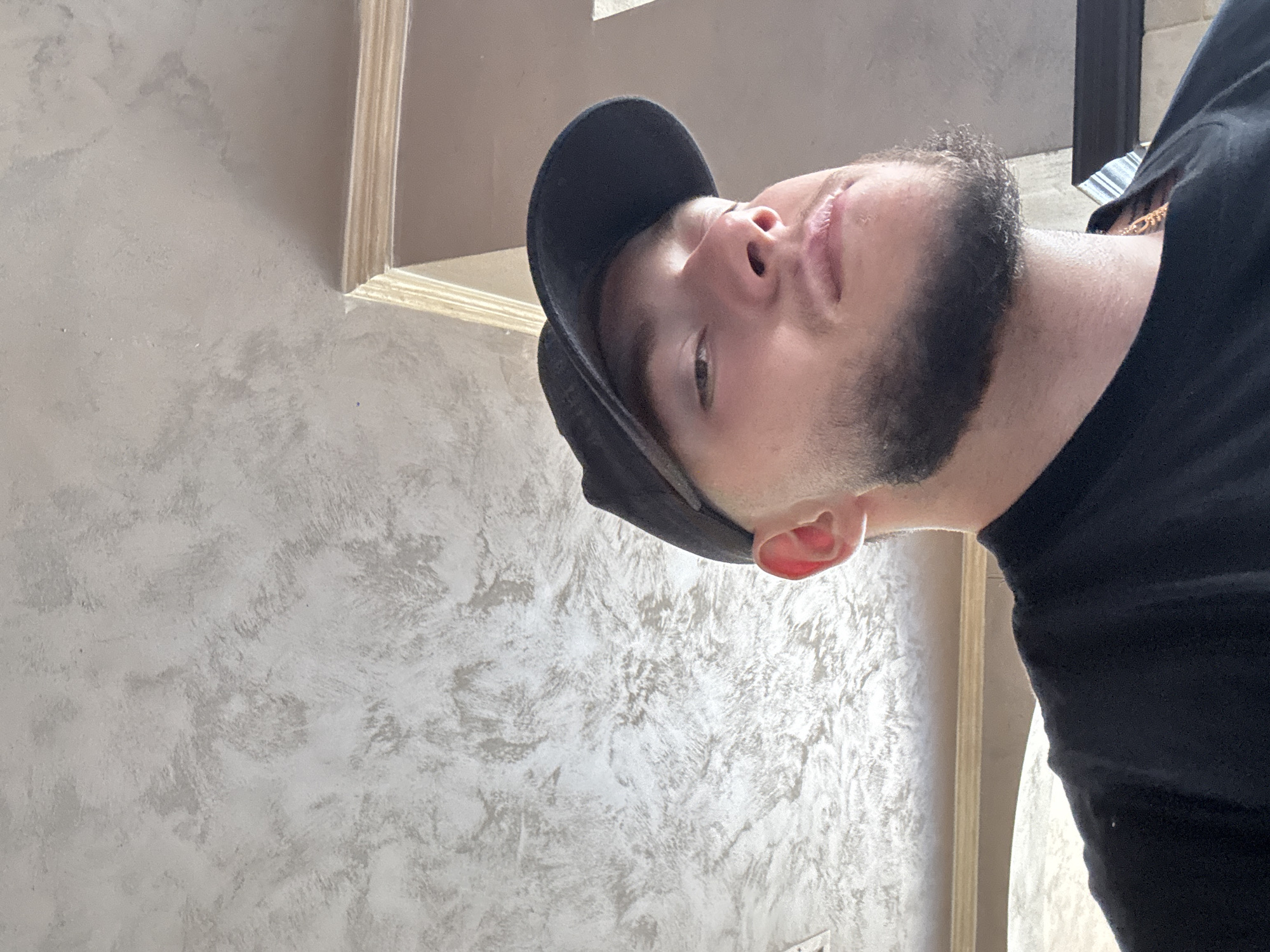 A low-angle medium shot shows a young man wearing a black baseball cap and a black t-shirt. He has a beard and is looking upwards towards the camera. The background is a textured ceiling and part of a wall with molding. There is no indication of the city of Nesebar, Bulgaria.