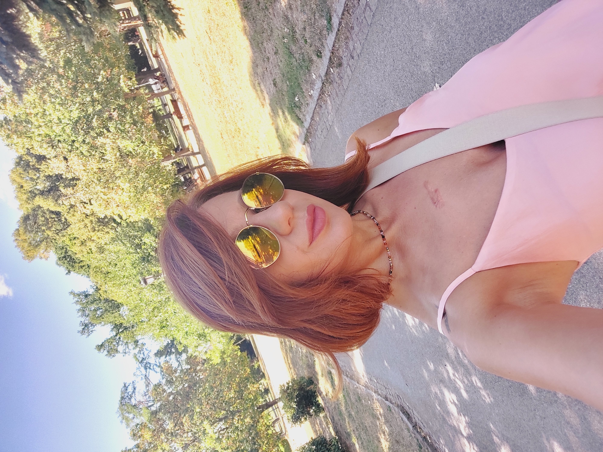 A woman with short, reddish-brown hair and sunglasses is taking a selfie in a park. She is wearing a pink dress and a necklace. There is a scar on her chest. The background shows trees and a path. The location is the Borisova Gradina park in Sofia, Bulgaria.