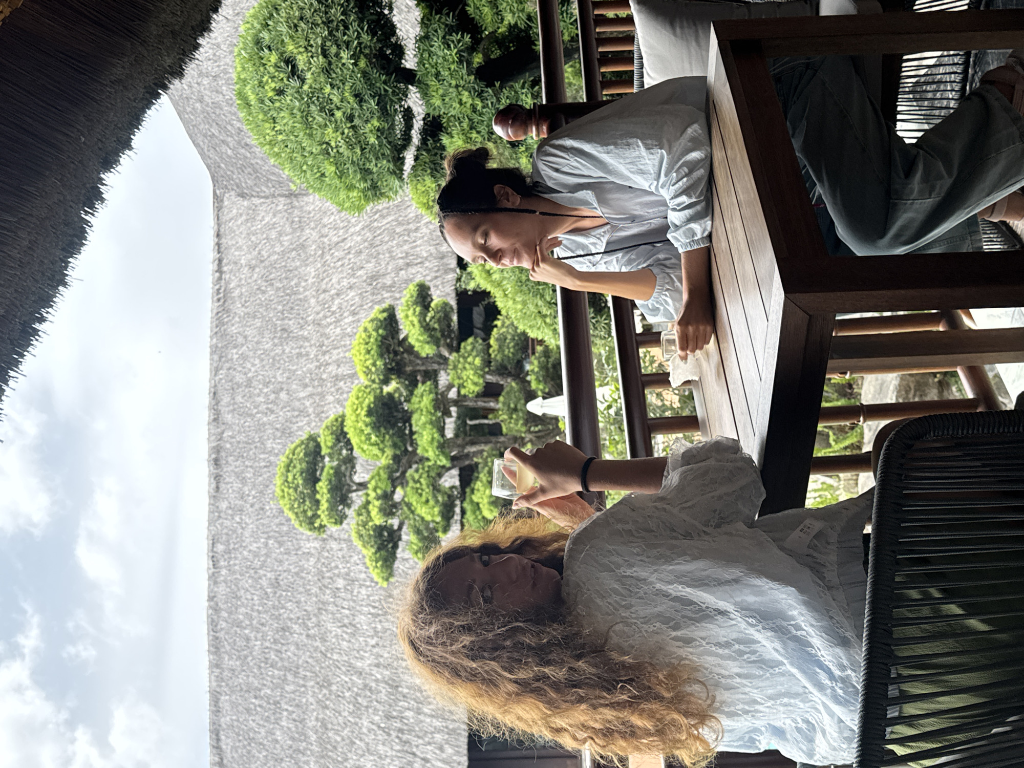 The image captures two women relaxing in an outdoor or semi-outdoor setting, possibly a resort balcony or lounge in Púng Luông, Vietnam.
In the foreground, on the left, a woman with long, curly light brown hair and a white lacy top holds a clear glass with a light-colored drink. Her gaze is directed slightly downwards. On the right, another woman, with dark hair tied back and wearing a light blue long-sleeved shirt, leans comfortably on a sturdy wooden table, resting her chin on her hand as she looks towards the first woman. Both women appear to be seated at the wooden table in woven chairs.
The background reveals a lush, verdant landscape with meticulously sculpted green trees, suggesting a well-maintained garden or natural area. A large, textured roof, possibly made of thatch or a similar natural material, is prominent on the left, hinting at traditional local architecture. The sky above, visible on the upper left, is bright with scattered clouds, indicating a daytime scene, likely late morning or early afternoon. The overall atmosphere conveys a sense of tranquility and leisure, with the women appearing relaxed and possibly engaged in conversation amidst the beautiful natural surroundings. No legible text is visible in the media file.