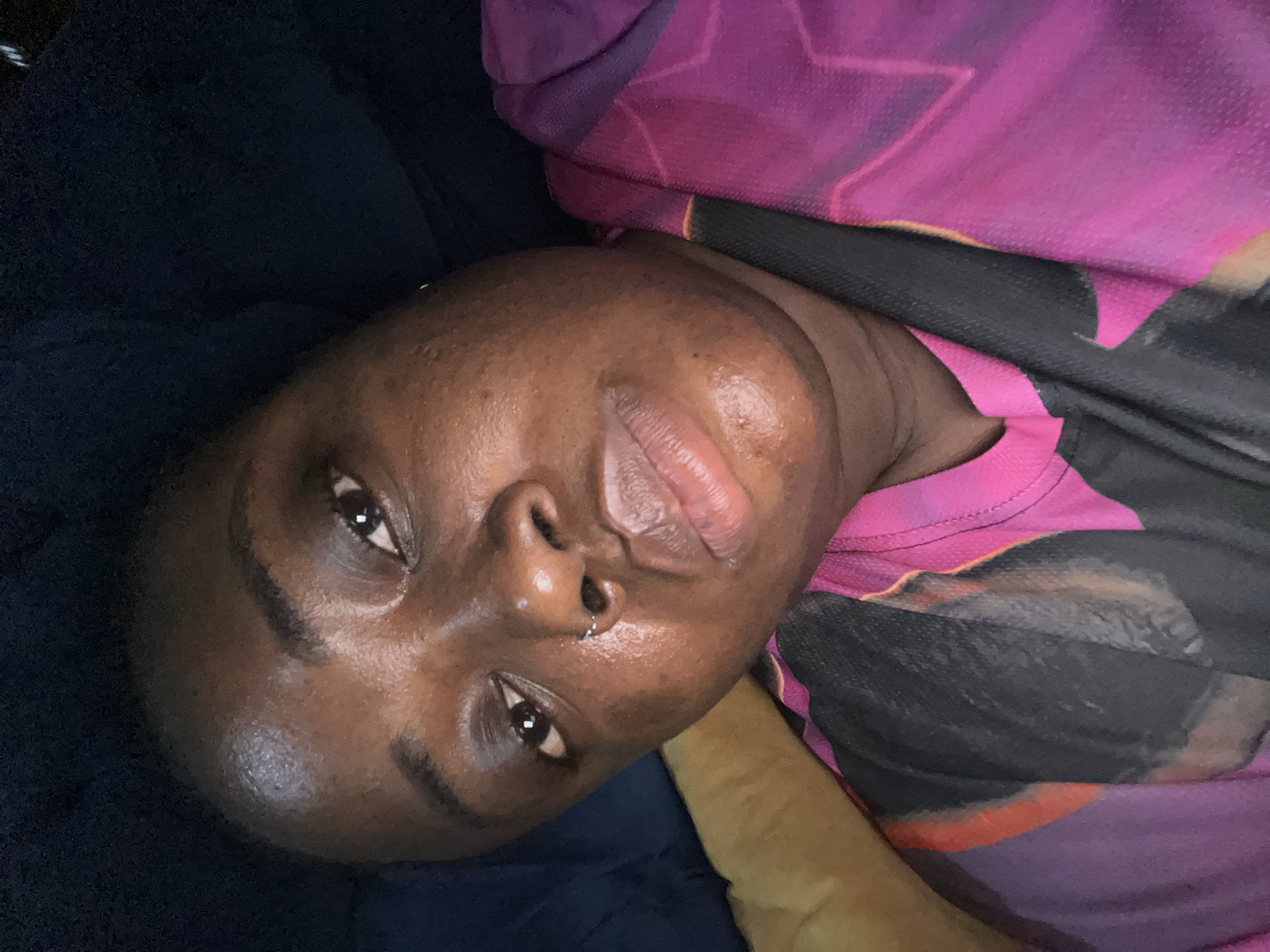 An individual with dark skin and dark eyes is lying down. They appear to be resting on a dark blue surface, possibly a bed or couch. They are wearing a pink and black top. A small, circular nose ring is visible in their left nostril. The photograph is taken at a close-up angle, focusing on the individual's face. The location in the city of Kubwa, Nigeria is not ascertainable from the image.