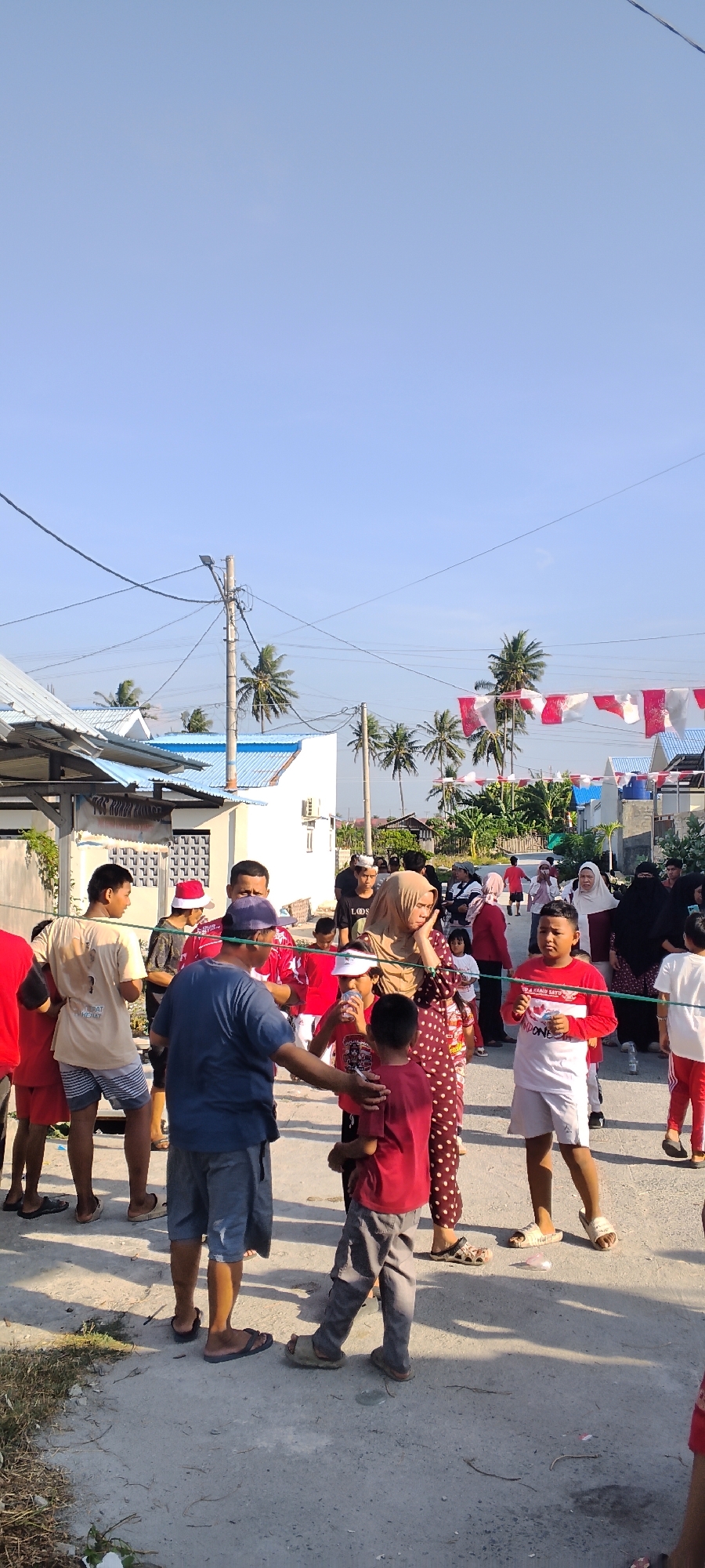 In Tawanjuka, Indonesia, a street is filled with people of various ages. The scene appears to be a community gathering, possibly a festival or celebration, given the presence of red and white flags strung across the street and on buildings. A clear blue sky looms above.
The individuals are dressed in casual attire, with many wearing red and white, suggesting a patriotic theme. Several children are present, some holding drinks. Adults are engaged in conversations, with one man in a blue shirt interacting with a younger boy in red. A woman in a patterned dress and a headscarf is also visible, her hand near her face, perhaps in thought or concern. Buildings with blue roofs line the street, and palm trees dot the landscape. The ground is paved with asphalt and concrete, with some dry vegetation and debris scattered along the edges. Power lines and a lamppost are also present, indicating a developed area within the city.