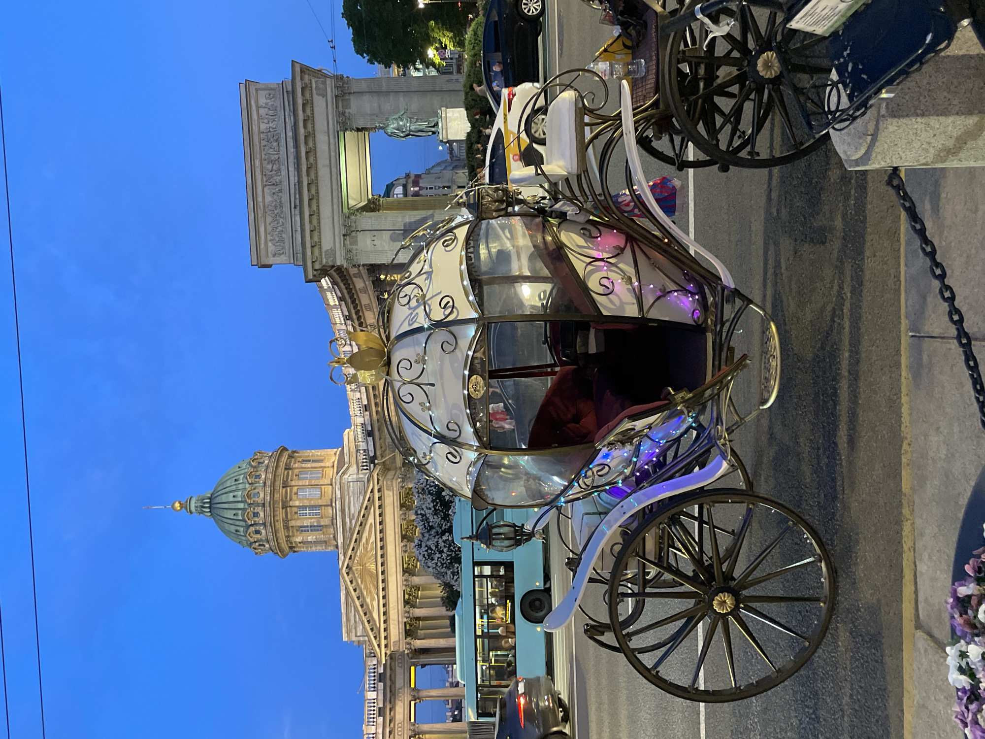 This image depicts a street scene in Saint Petersburg, Russia, at twilight, with the Kazan Cathedral and the General Staff Arch prominent in the background.
In the foreground, a highly decorated, Cinderella-style pedicab or carriage occupies the left-center. It features a white frame with gold scrollwork accents and a clear, domed canopy. The carriage is illuminated by multi-colored LED string lights (pink, purple, blue) and contains a dark red upholstered interior. It rests on large, black, spoked wheels with gold central hubs. To the right of the carriage, a small, cushioned white seat is visible, along with a plastic water bottle placed on a small shelf. The immediate bottom foreground reveals part of a dark planter containing purple and white flowers, and a concrete barrier with a metal chain.
The midground features a paved street with several vehicles. A light blue city bus, filled with passengers, is visible on the left, moving away from the viewer. A dark grey sedan is partially visible ahead of the bus. To the right, a white taxi with yellow markings and a black sedan are present behind the carriage, also on the street.
The background is dominated by significant architectural landmarks. On the left, the illuminated Kazan Cathedral stands with its distinctive large green dome topped with a golden finial. To the right of the cathedral, a large, illuminated classical archway (part of the General Staff Building) spans an opening. Through this archway, a statue of a man on a pedestal (Field Marshal Mikhail Kutuzov) is visible. Further to the far right, illuminated green trees and other structures can be discerned. The sky is a deep, clear twilight blue.