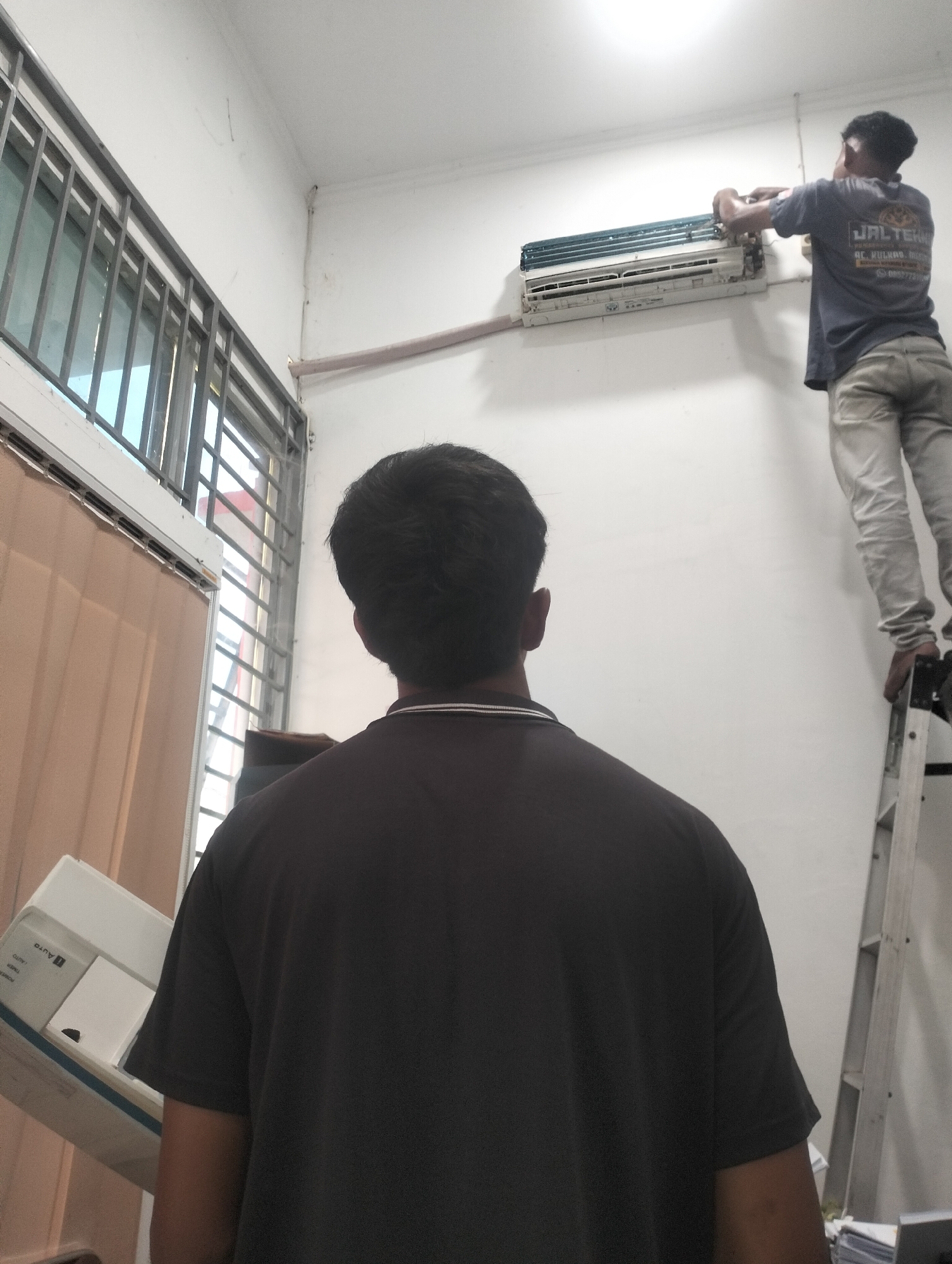 In an indoor environment, likely a commercial or residential building in Lhokseumawe, Indonesia, two men are present. One man, viewed from behind and closer to the foreground, wears a dark t-shirt. He appears to be observing the activity of the second man. The second man, positioned further in the background and higher up, is standing on a silver ladder. He is actively working on a wall-mounted air conditioning unit, reaching towards it with both hands. He is wearing a dark t-shirt with a logo and text, and light-colored pants. The air conditioning unit is white and partially disassembled or being serviced. The walls are painted white, and a window with metal bars and a beige curtain is visible on the left side of the image. A cream-colored pipe runs horizontally along the wall near the air conditioner. The overall scene depicts a repair or installation of an air conditioning unit.