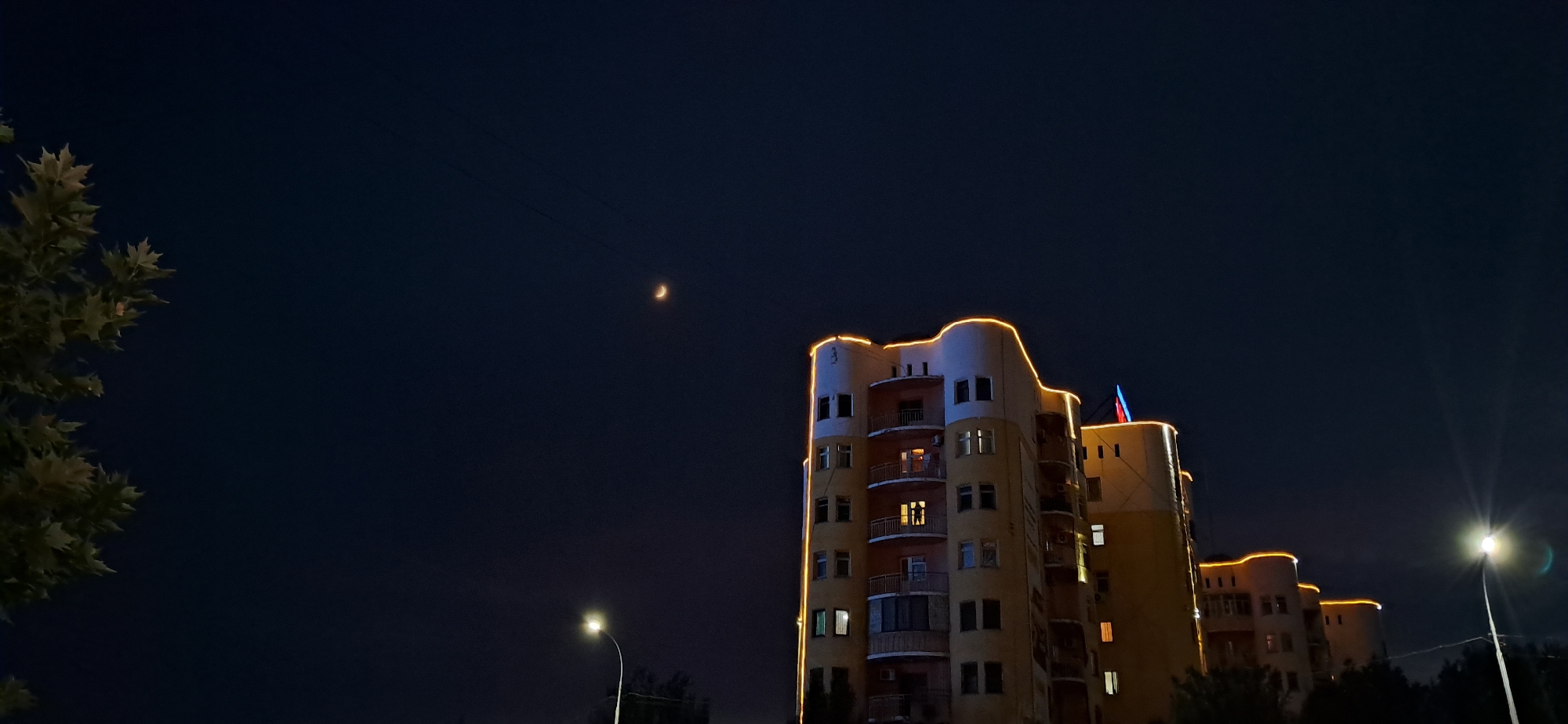An eye-level, medium outdoor shot captures a city scene at night under a dark blue sky. On the left, the dark green foliage of a tree occupies the left third of the frame. The center and right feature a multi-story, light yellow building with rounded balconies. The building is illuminated by a string of warm yellow lights outlining its upper floors and a Russian flag positioned on the top right. A thin, crescent moon is visible in the upper center of the sky, with power lines running horizontally across the frame. In the lower center, a streetlamp with a bright white light shines upwards, and the lower right shows a bright streetlamp with lens flare. Trees are visible at the base of the building and in the lower right corner.