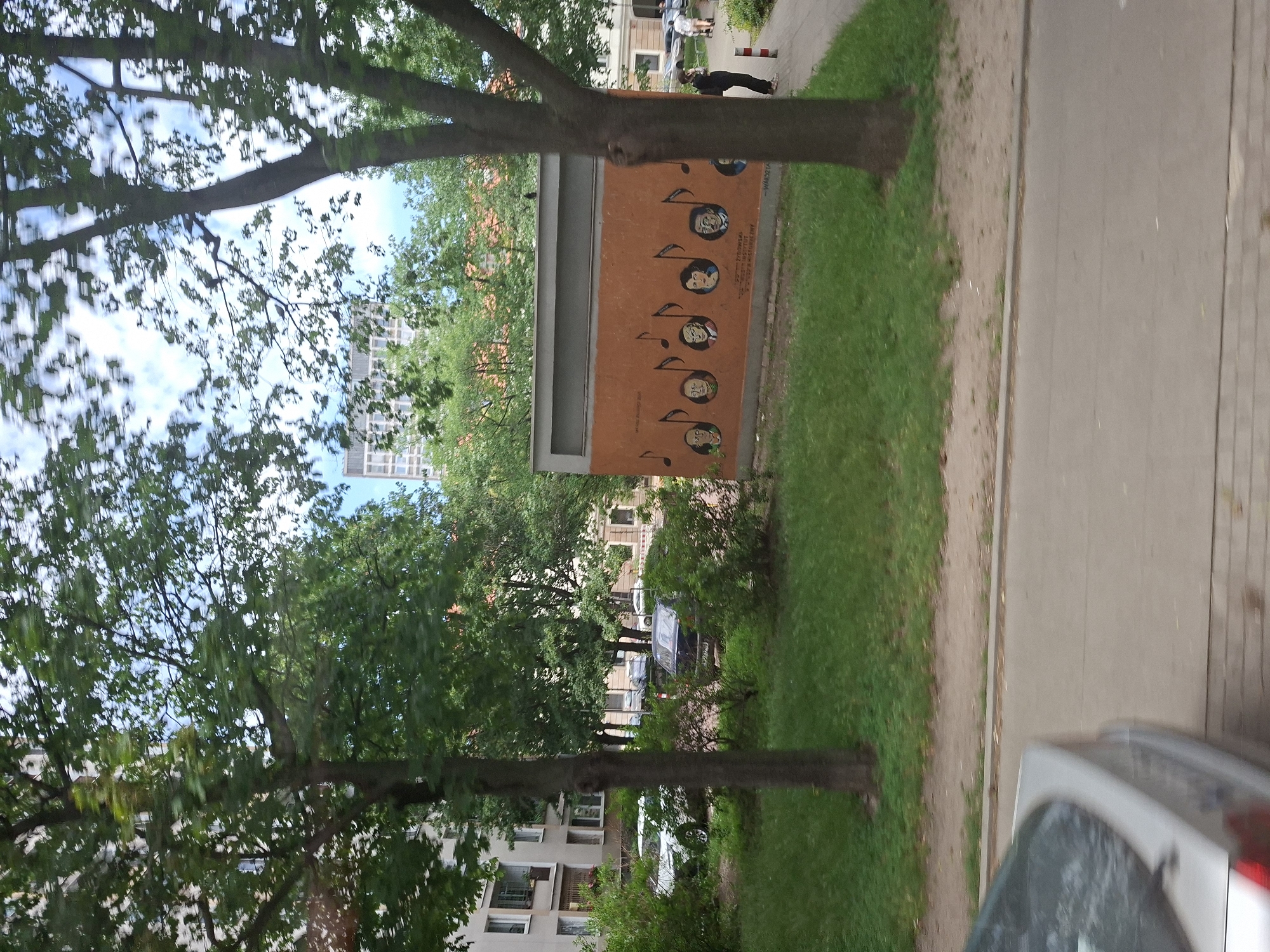 The image shows an outdoor scene with trees, buildings, and a sidewalk. In the foreground, there are several large trees with green leaves, suggesting a warm season. One tree trunk is prominent in the center. Behind the trees, there are apartment buildings with visible windows and balconies.
On an orange panel mounted on a structure, there is artwork depicting several circular portraits of people, arranged vertically and accompanied by musical notes. The portraits appear to be in a cartoonish style. Some text is visible on the orange panel, but it is not clearly legible.
To the right of the panel, there is a strip of green grass and a sandy path, leading to a paved sidewalk or road. A silver car is partially visible in the lower right corner of the image, and a dark car is parked further down the street.
In the background, there are more buildings and trees. A few people are visible walking along the sidewalk, one of whom is wearing dark clothing and appears to be looking down. The sky is partially visible and appears cloudy. The overall impression is a pleasant day in an urban environment, likely a residential area. The presence of the musical notes and portraits suggests a cultural or artistic element within the setting. The orientation of the image is sideways.