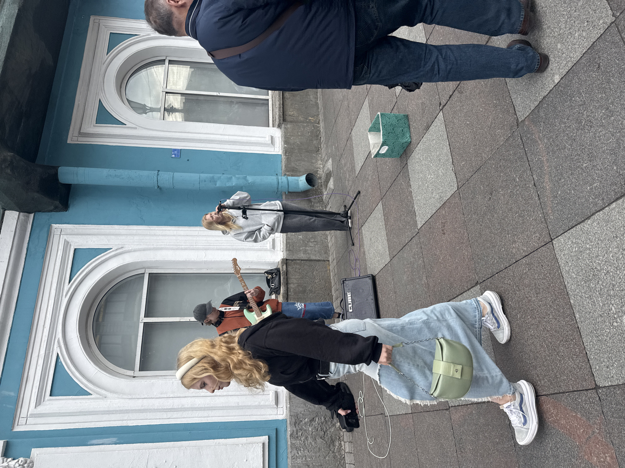 The image depicts an outdoor street performance in Saint Petersburg, Russia. In the foreground, a young woman with blonde hair, wearing a black hoodie, a distressed denim skirt, and white sneakers, walks from left to right, carrying a green handbag and a black electronic device. Behind her, a band performs on the sidewalk in front of a bright blue building with arched windows. A male guitarist in an orange vest and baseball cap is positioned to the left, while a female vocalist stands at a microphone to the right. An amplifier is visible on the ground near the guitarist. A green patterned box, possibly for donations, sits on the sidewalk to the right of the performers. A man in a dark jacket and jeans stands facing away from the camera on the far right, observing the scene. The architectural style of the building suggests it is located in the historic center of Saint Petersburg.