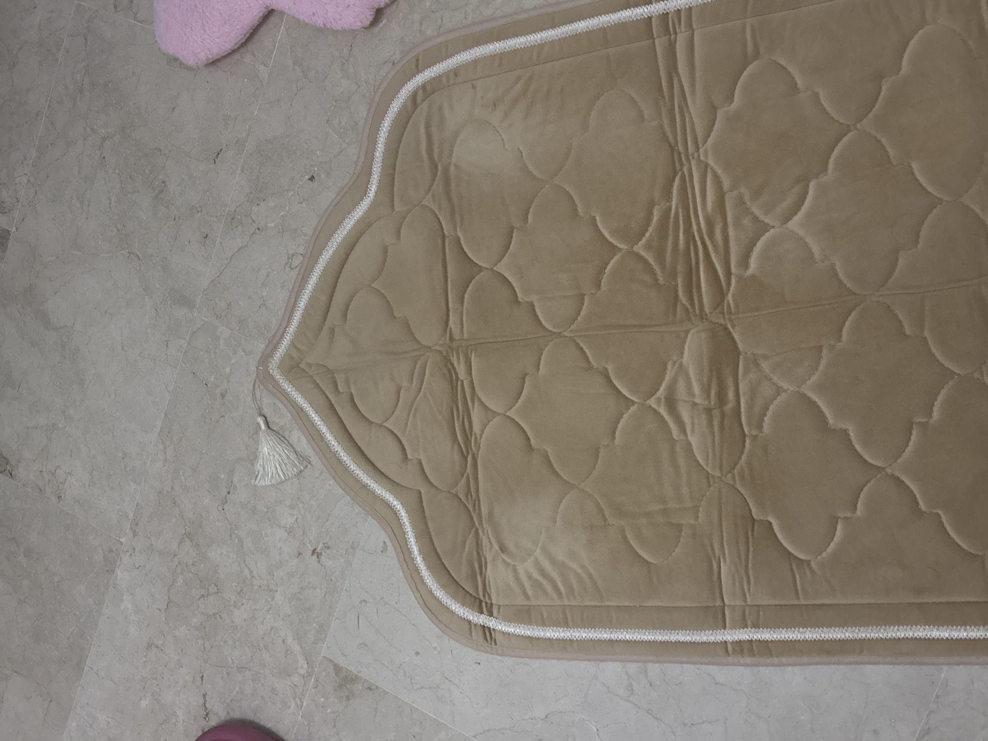 The image shows a close-up, high-angle view of a beige prayer rug with a decorative quilted pattern on a light-colored tiled floor. A white tassel hangs from the top left corner of the rug. The rug has a double white trim along its curved edges. To the right of the rug, a blurry pink fuzzy object is partially visible. There is no information in the image to suggest the location in the city of Orihuela, Spain, or any specific actions or interactions.