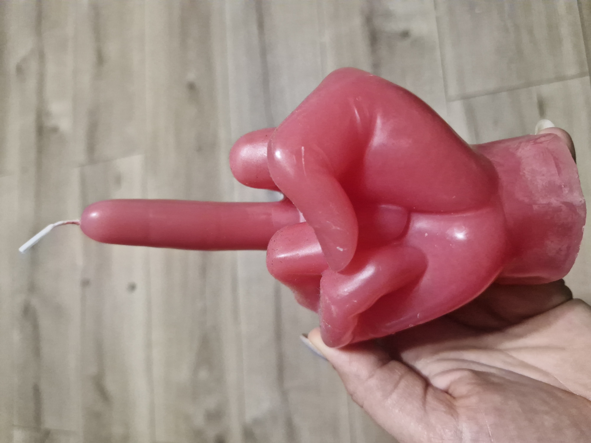 A pink, fist-shaped candle, giving the middle finger, is being held in a person's hand. The background is a light brown, wood-patterned surface. There is no indication of the city of Plovdiv, Bulgaria.