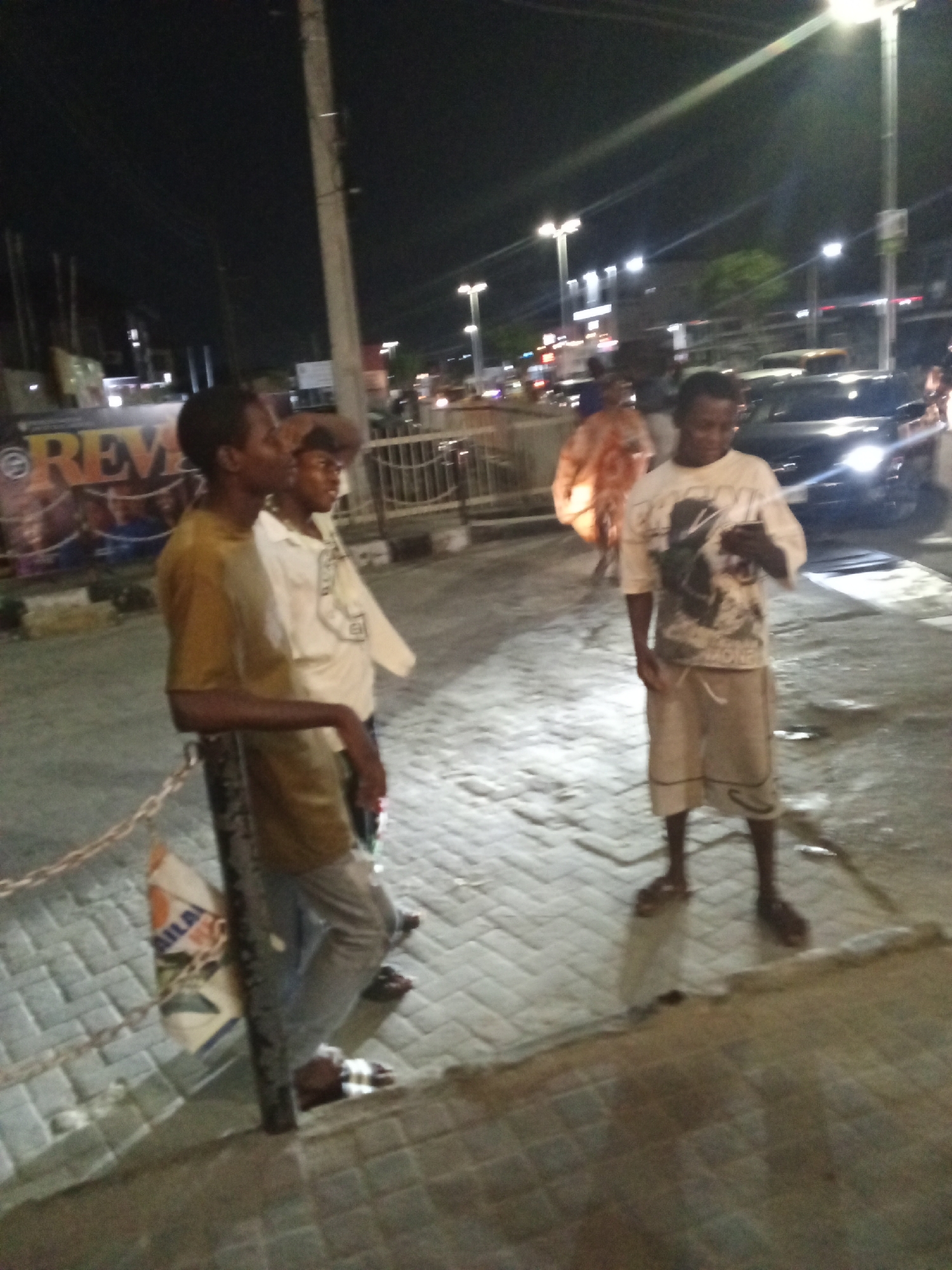The image displays a nighttime street scene, likely in an urban or semi-urban area. The primary subjects are three men standing on what appears to be a paved sidewalk or public space.
On the far left, a man wearing a mustard-yellow short-sleeved shirt and light-colored trousers leans casually against a dark, sturdy pole. A white and orange bag with partially visible text "AILA" hangs near the base of the pole. Slightly behind him and to his right, another man in a light-colored collared shirt looks towards the right.
Further to the right, a third man stands centrally, wearing a light-colored T-shirt with a dark graphic print and knee-length shorts. He is looking down at a small object held in his hands, likely a mobile phone. All three men appear to be wearing open-toed footwear like sandals.
The ground is paved with light-colored interlocking bricks. The scene is illuminated by artificial light sources, including multiple bright streetlights in the background that create noticeable lens flares. Vehicles with illuminated headlights, including a dark car on the right, are visible on the street beyond the individuals. A large banner or sign in the left background prominently displays the orange letters "REV" against a dark pattern. The overall activity suggests people casually standing or conversing on a street at night.
