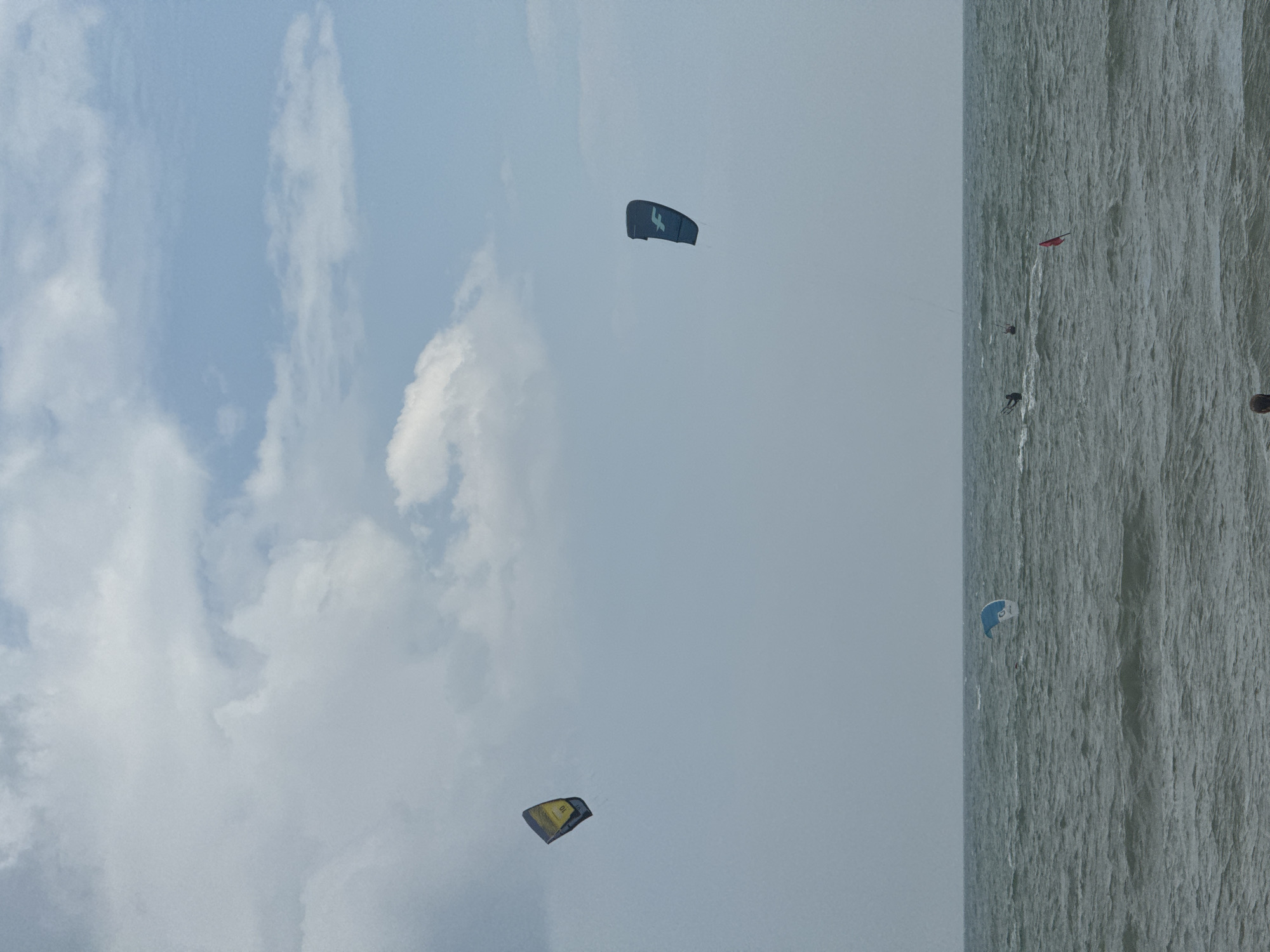 The image depicts a bright, overcast day at Mui Ne Beach, Vietnam, with the turquoise ocean dominating the lower half of the frame. The sea is choppy, with moderate waves creating whitecaps. Two kites, one yellow and black with the number "10" visible and the other dark blue with a white logo, are prominently displayed in the sky. Several figures are engaged in kitesurfing on the water, appearing as small dark shapes against the waves. A small red flag is also visible on the water's surface. The sky is a pale blue, interspersed with large, fluffy white clouds, suggesting a humid, possibly tropical, atmosphere. In the foreground, a blurred silhouette of a person's head and shoulders is visible, suggesting the photographer is standing on the shore.