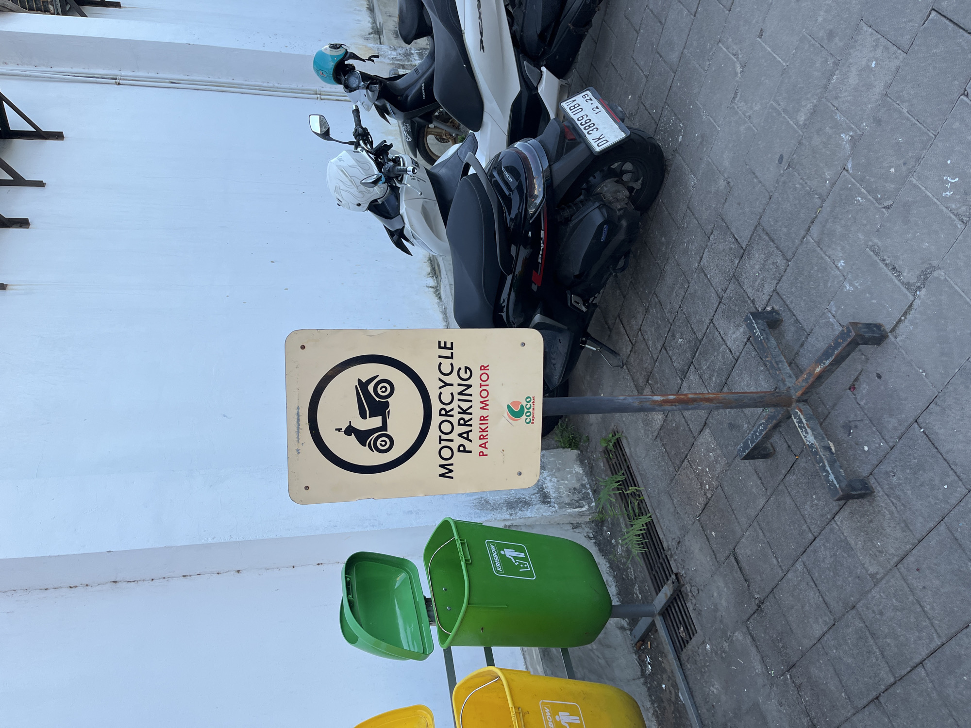 The image shows a motorcycle parking area in an urban setting in an unknown city in Indonesia. Two motorcycles are parked beside a white building wall. A sign indicates the area is designated for "Motorcycle Parking" or "Parkir Motor" in Indonesian, with the Coco Supermarket logo. A metal stand holds the sign and a stack of waste bins. The waste bins are colored green, yellow, and another color is seen under the yellow bin. The bins each have a different waste sorting symbol on them. The ground is paved with interlocking gray bricks. The motorcycles partially obscure a ground-level drain covered with a metal grate. The license plate on the motorbike reads "DK 3869 UBV".