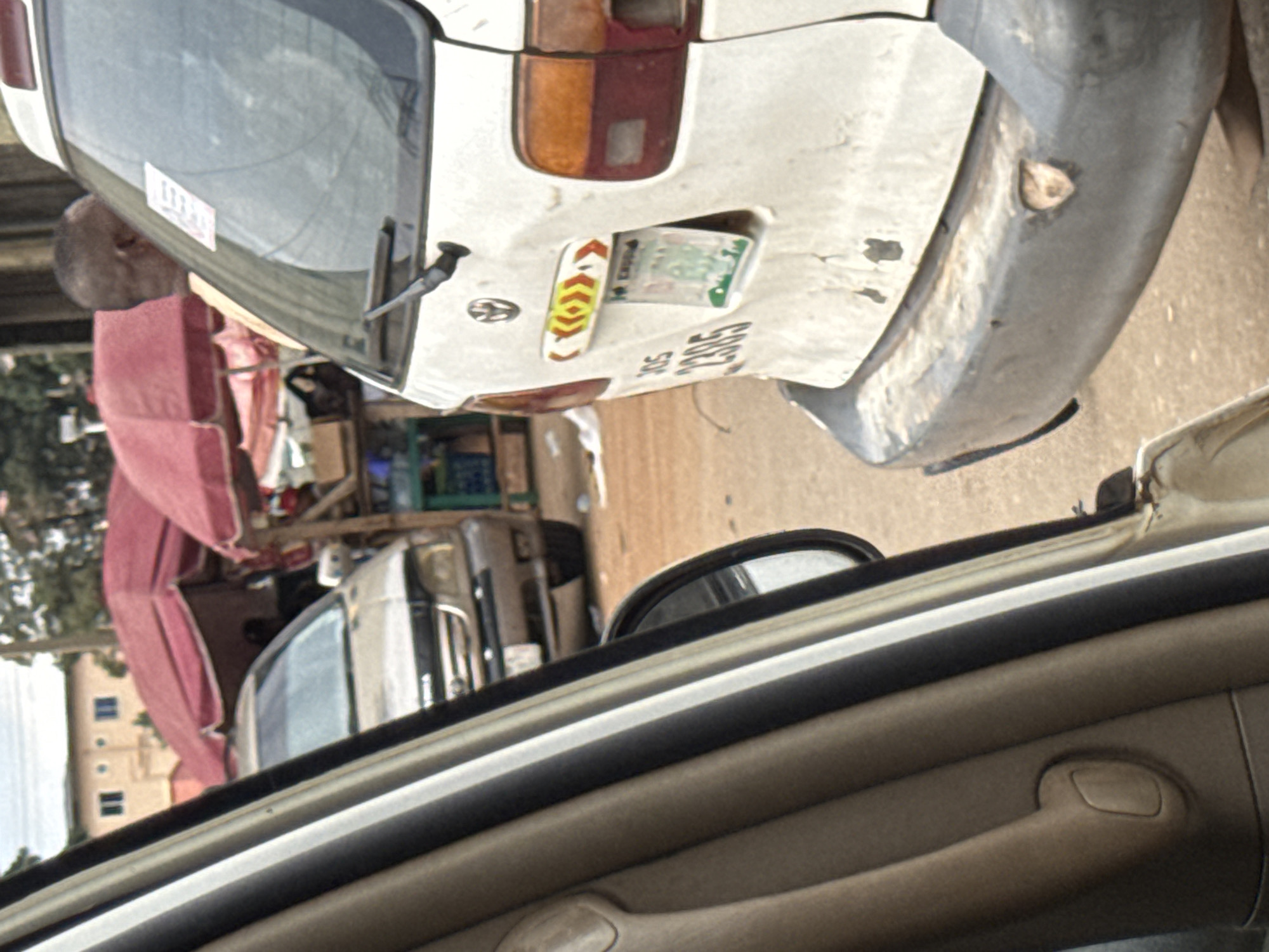 The image depicts a street scene in Orji, Nigeria, likely captured from within a vehicle. In the foreground, the interior of a car is visible on the left, with beige and black trim. Outside the car, a white van is parked, its rear end prominently featured. The van has a dented bumper and a license plate with the numbers "2395". A colorful decal with red, yellow, and white arrows is above the license plate. To the left of the van, another car, possibly a silver SUV, is partially visible. In the background, market stalls with red awnings are set up, suggesting a bustling commercial area. A person with dark skin and short hair, wearing an orange shirt, is standing near the rear of the white van, looking away from the camera. The ground is a dusty, reddish-brown surface. The overall impression is of a typical urban scene with elements of commerce and transportation.