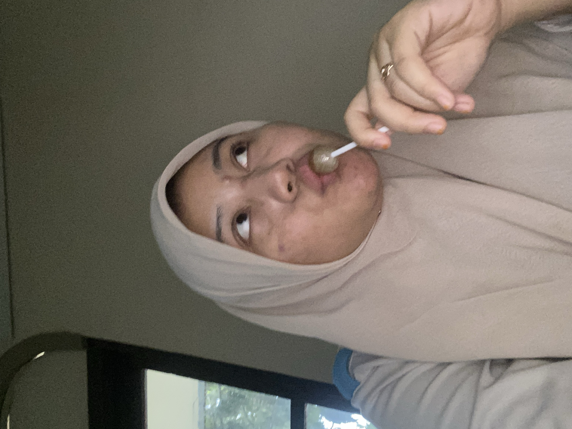 A woman wearing a beige hijab and light-colored shirt is in a close-up shot. She is holding a lollipop to her lips and looking upwards. A ring is visible on her right hand. The background is a plain, textured wall. The scene appears to be indoors, possibly in a residence.