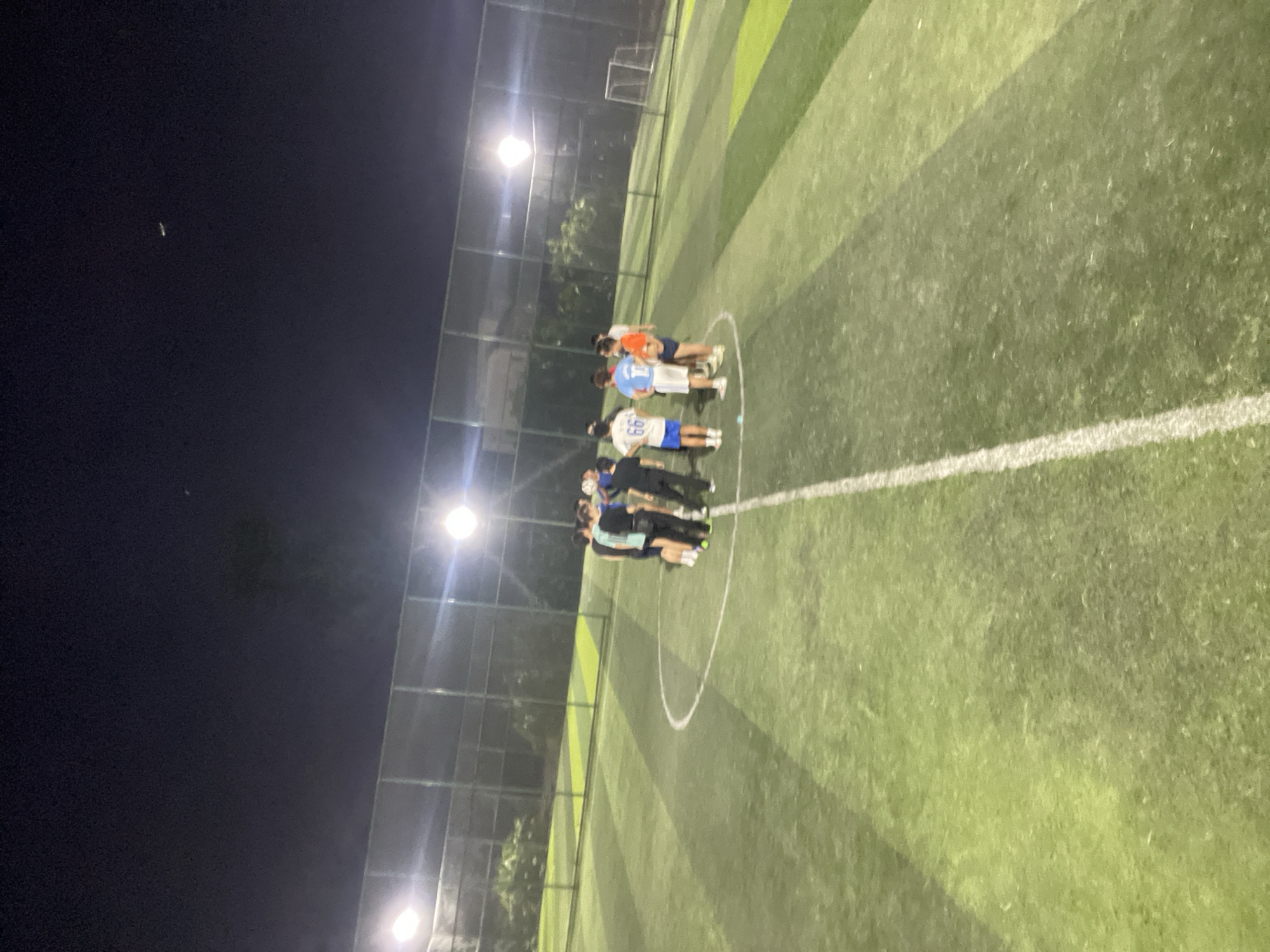 A group of people stand in a circle on a green artificial turf soccer field at night. The field is illuminated by bright lights along a fence. The location is likely Jiangjin, China. A soccer goal is visible in the background. The people seem to be interacting or talking. The sky is dark.