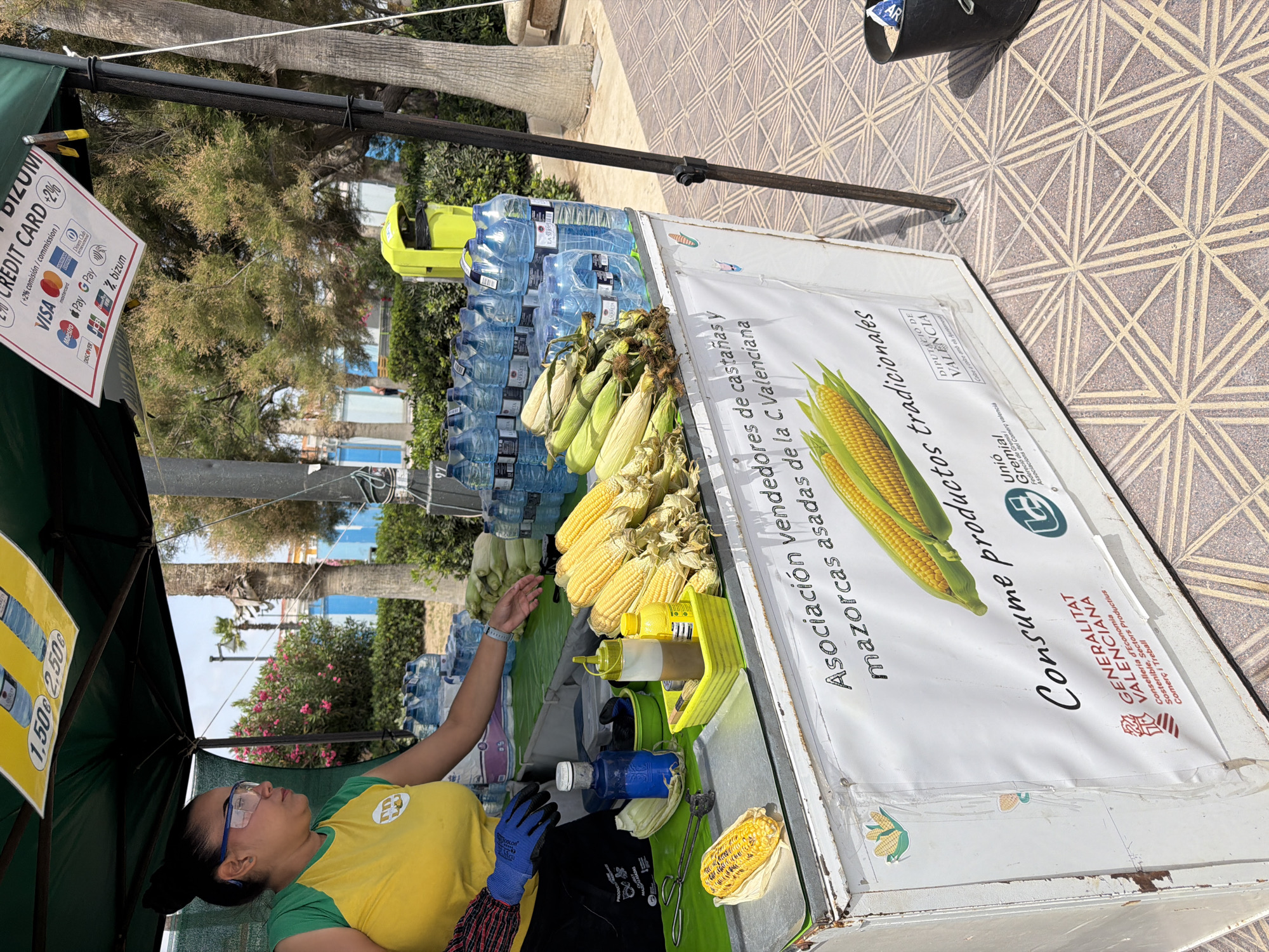 In an outdoor setting, a woman wearing a yellow and green shirt and blue protective glasses is standing behind a white food cart. She has dark hair pulled back and is wearing blue gloves. In front of her on the cart, there are several ears of corn, some shucked and some with husks. There are also bottles of water and condiments. A sign on the cart reads "Asociación vendedores de castañas y mazorcas asadas de la C. Valenciana Consume productos tradicionales". Another sign above the cart displays prices and accepted payment methods, including credit cards and Bitcoin. The cart is set up on a tiled pavement, with trees and buildings visible in the background.