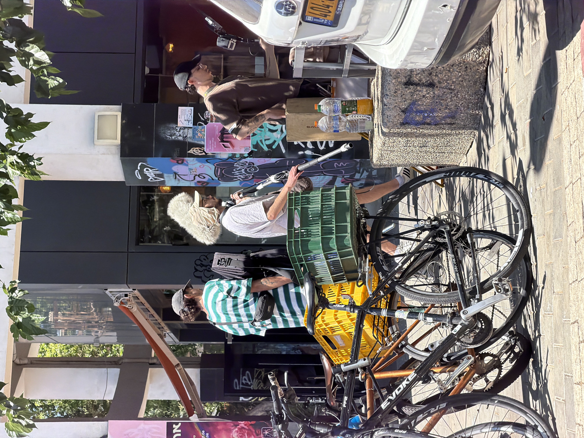This photograph captures a street scene in Tel-Aviv, Israel, on a sunny day. In the foreground, several bicycles are parked on a brick-paved sidewalk. One bicycle, in the center, is black and has a large yellow plastic crate attached to its rear rack. To its left, another bicycle is partially visible, appearing to be orange and black. Further left, a third bicycle's front wheel is in the frame.
In the middle ground, three individuals are present. On the left, a person wearing a green and white striped t-shirt and a black fanny pack stands facing away from the camera. Next to them, another person is holding a long, thin object, possibly a microphone boom, and wearing a light-colored hat with a furry trim. To the right of these two, a third person, wearing a brown t-shirt and a black baseball cap, is looking down and appears to be interacting with something out of frame. Behind this group, there is a building with dark gray siding and colorful graffiti. A white Subaru car is parked on the right side of the image, its rear bumper and license plate visible. Two plastic water bottles and a juice bottle sit on a concrete block near the right side of the frame. The scene suggests a casual outdoor gathering or event.