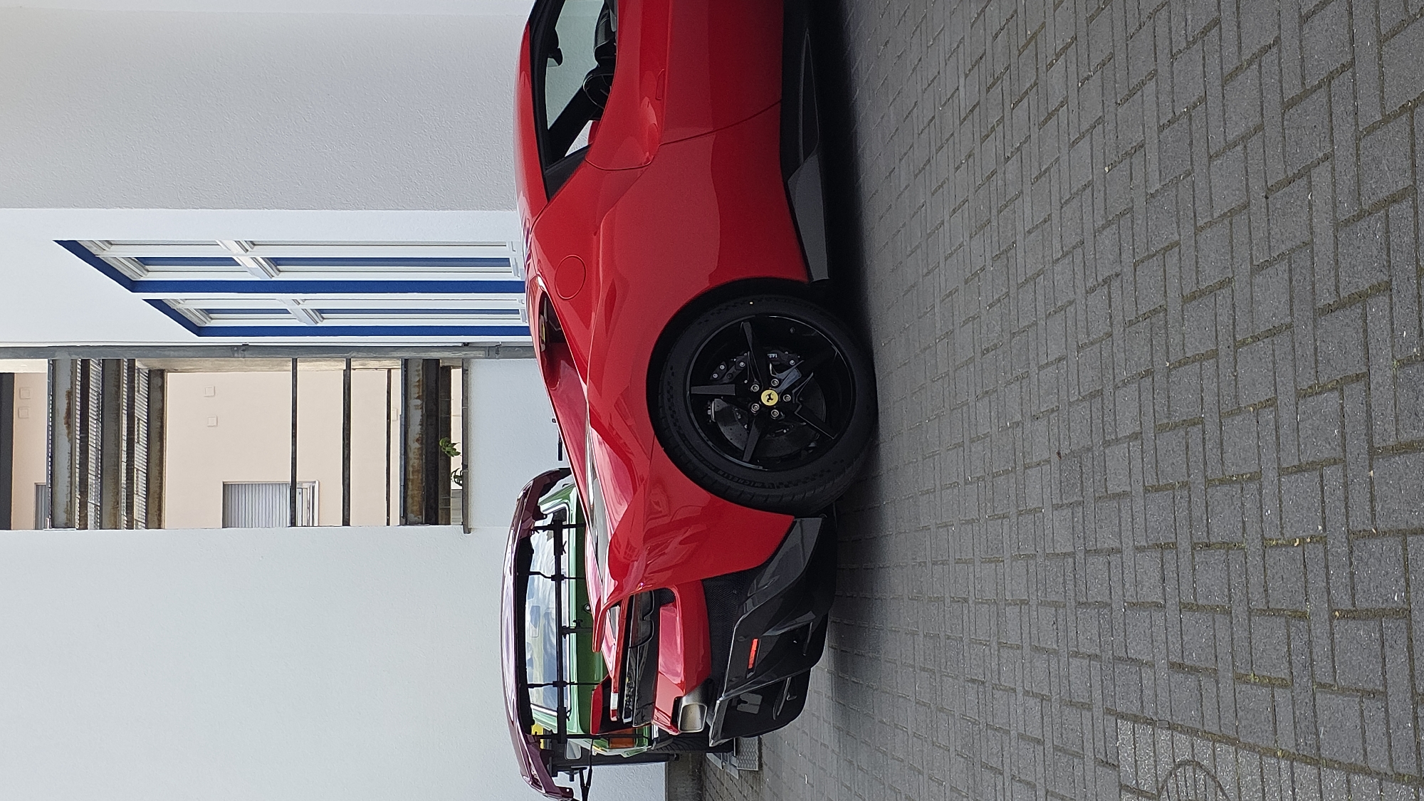 A red sports car is parked on a paved surface, viewed from the rear quarter. The car has a low profile, with prominent black accents on its rear bumper and diffuser. It features a black wheel with a five-spoke design and a yellow Ferrari emblem at its center. To the left of the red car, partially visible, is a green vintage car with a dark roof rack. Behind the cars, a building with white walls and blue-trimmed windows is visible. The background also shows another building with light-colored walls and multiple balconies or external staircases. The ground is covered with gray brick pavers arranged in a patterned layout.