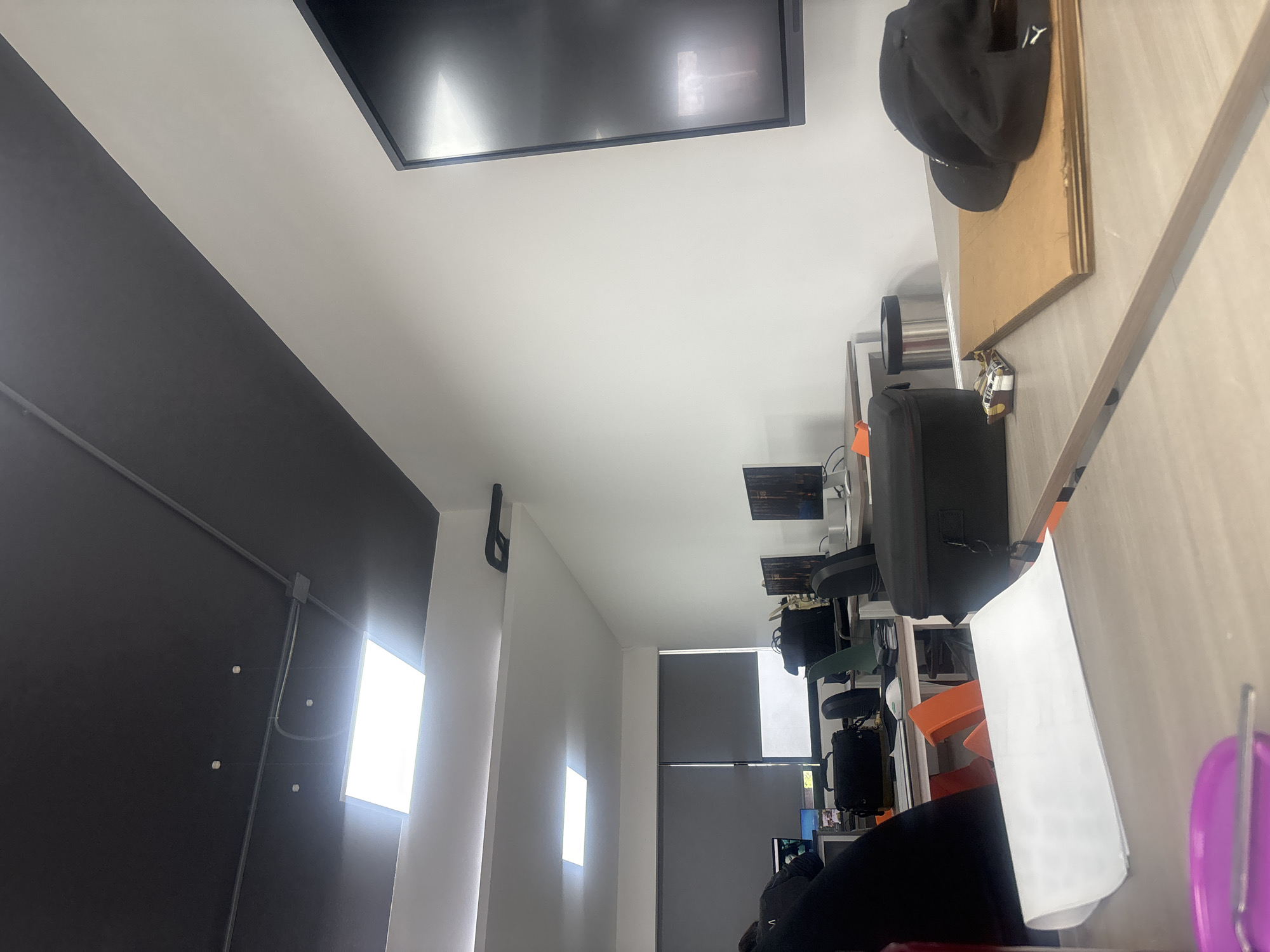 The image shows the interior of what appears to be an office space. The walls are painted in a two-tone design: the lower portion is dark grey, while the upper portion is white. There are illuminated rectangular light fixtures mounted on the dark grey wall. A large, rectangular flat-screen television is mounted on the white ceiling. A desk or countertop runs along the right side of the frame, cluttered with objects including a black baseball cap, a backpack, and assorted office supplies. A window with grey blinds can be seen in the background. The scene takes place in the city of San Andrés Cholula, Mexico.