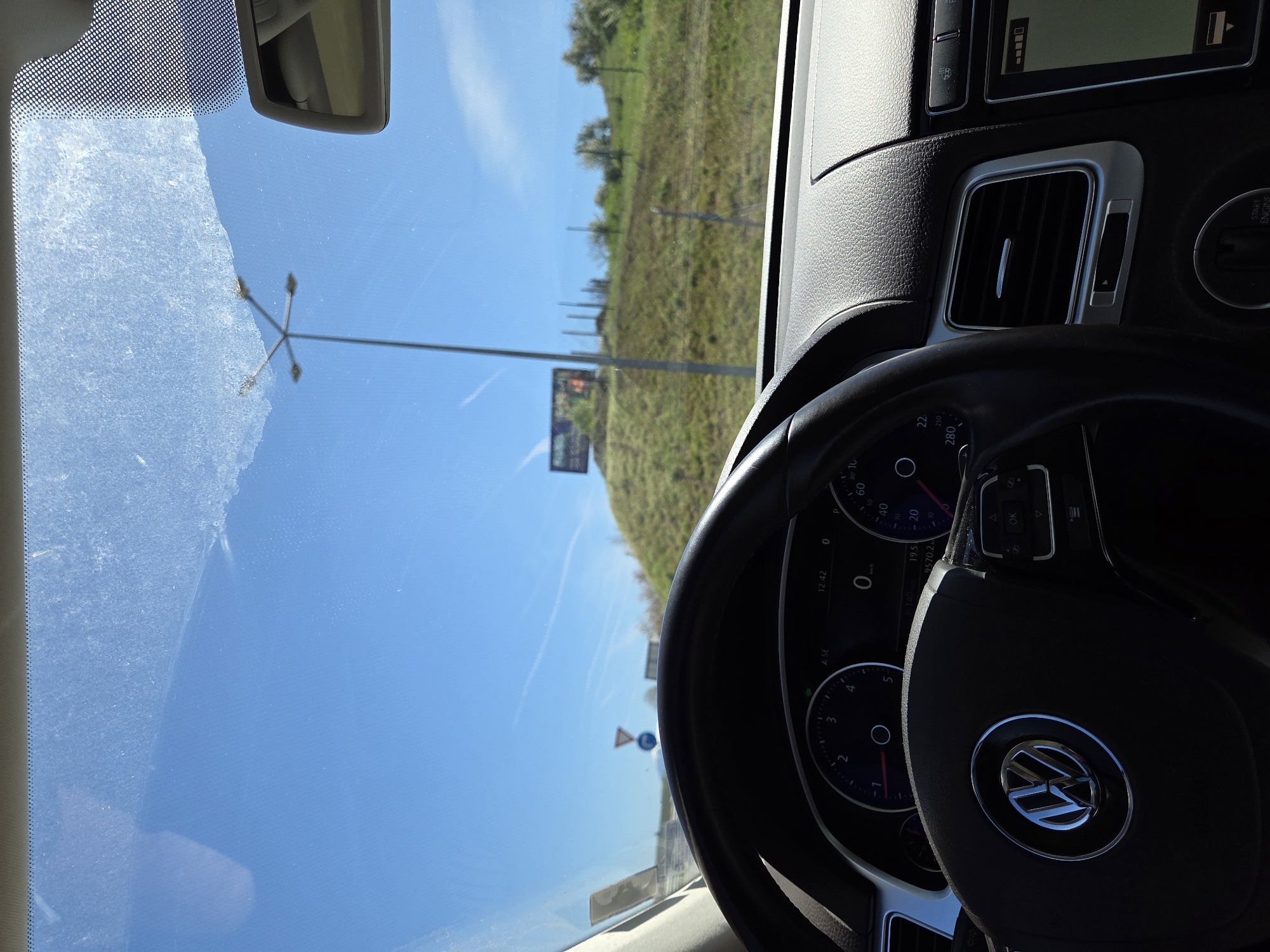 The image depicts the interior of a vehicle, likely a car, with the dashboard and steering wheel prominently visible. The steering wheel features a Volkswagen logo. Through the windshield, the external environment can be observed, showcasing a green, grassy landscape under a bright blue sky. Road signs and utility poles are also visible. Based on the OCR data present, it can be confirmed that the car is located in Levunovo, Bulgaria.