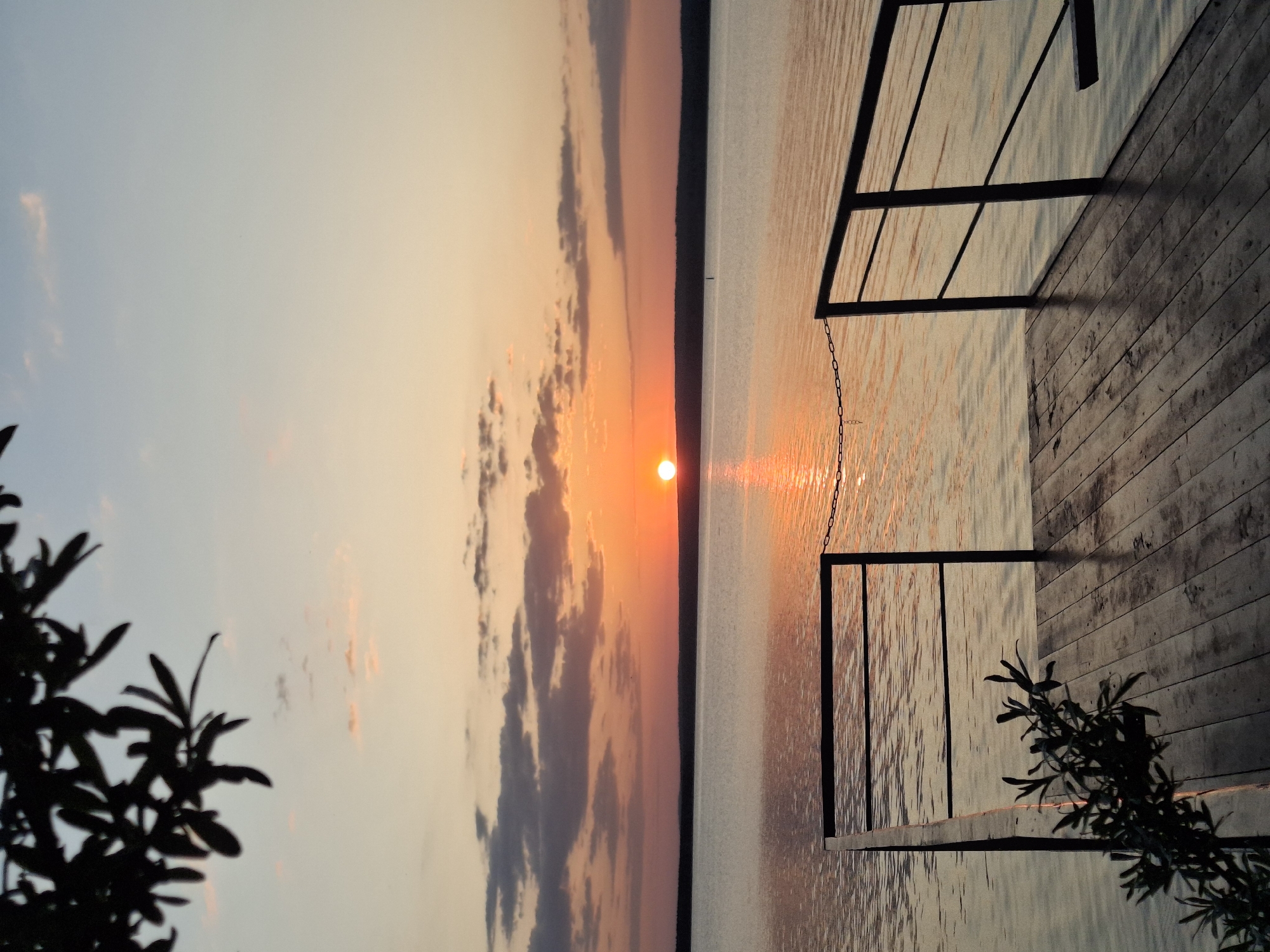 The image shows a sunset over a body of water. The sun is setting on the horizon, with clouds reflecting the orange and pink light. To the right of the frame, a wooden dock with a black metal railing extends into the water. A chain hangs from the railing into the water. Silhouetted foliage is visible in the bottom corners of the image. The location is likely near a body of water in or near Chaikovsky, Russia.