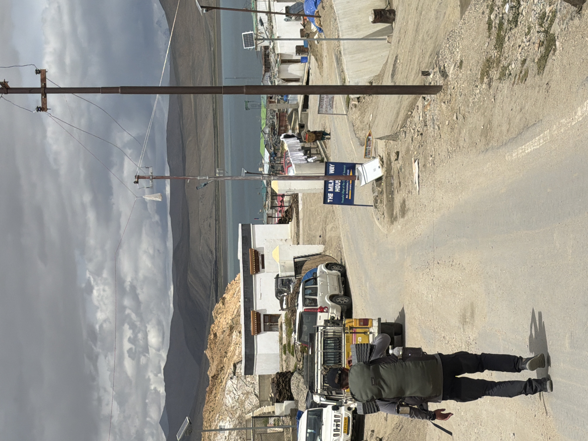 A man with a large backpack walks away from the camera on a dusty road in Karzok, India. He is wearing a gray long-sleeved shirt and black pants. In the background, several vehicles are parked, including a white SUV and a yellow pickup truck. A white building with a red-trimmed roof is visible behind the vehicles, and beyond that, a large body of water stretches towards distant, hazy mountains under a cloudy sky. A utility pole with power lines stands to the right of the frame. A sign that reads "THE MILK WAY HOUSE" is on the left side of the road. The environment appears arid and mountainous.