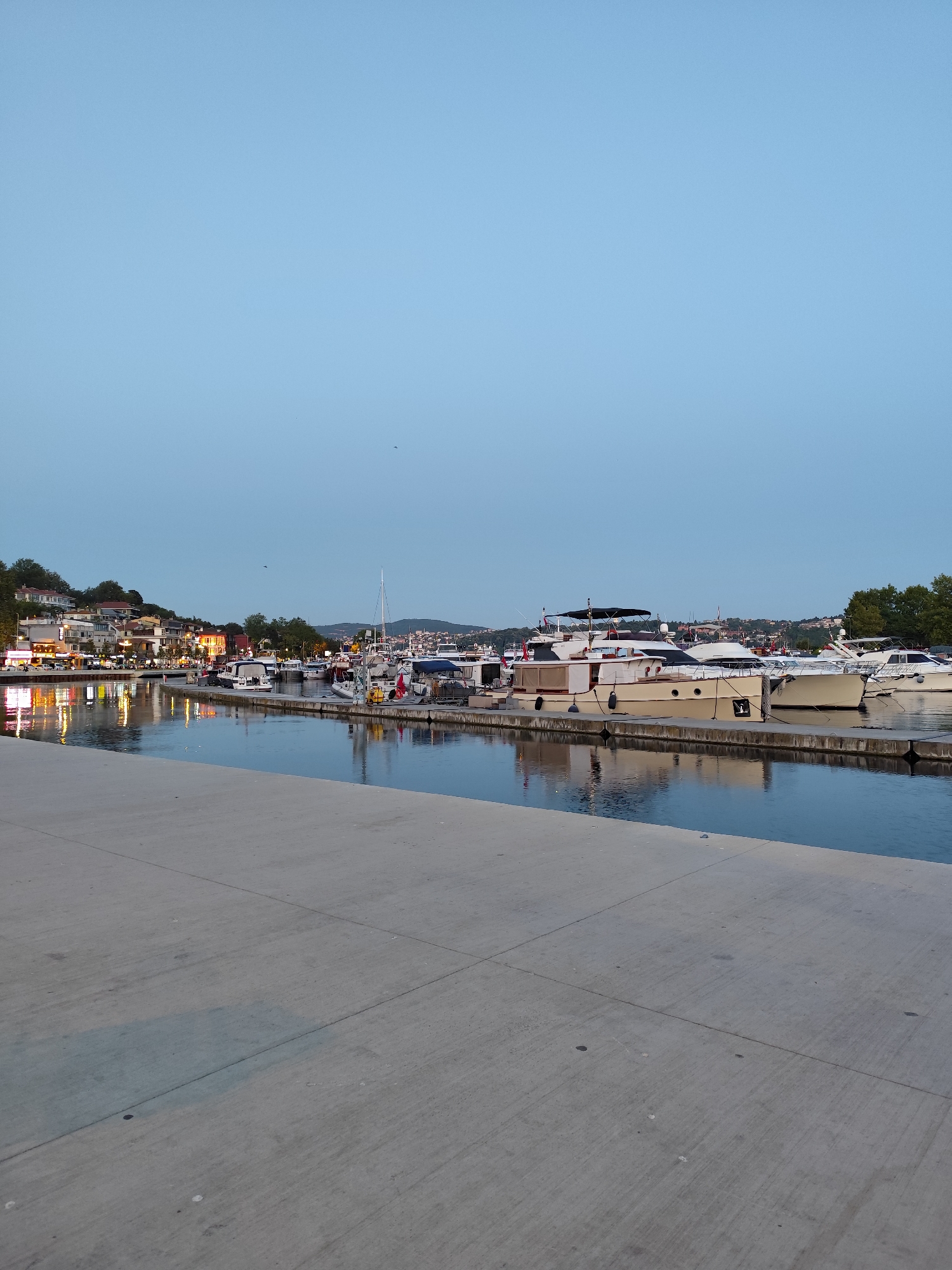 The image shows a view of a harbor in Sarıyer, Turkey, likely during the evening due to the ambient light. In the foreground, a concrete walkway runs along the edge of the water. The water is calm and reflects the lights from the buildings and boats.
A variety of boats are docked in the harbor, ranging from smaller pleasure crafts to larger yachts. Some boats are illuminated, casting warm glows on the water. In the background, a hillside is visible, dotted with buildings that appear to be homes and businesses. The buildings are illuminated, suggesting activity. Trees and greenery are interspersed along the hillside.
The sky is a clear, pale blue, indicating either early evening or a very clear twilight. A few distant birds are visible in the sky. The overall scene depicts a peaceful and picturesque harbor setting within the city of Sarıyer. There are no people clearly visible in the image.