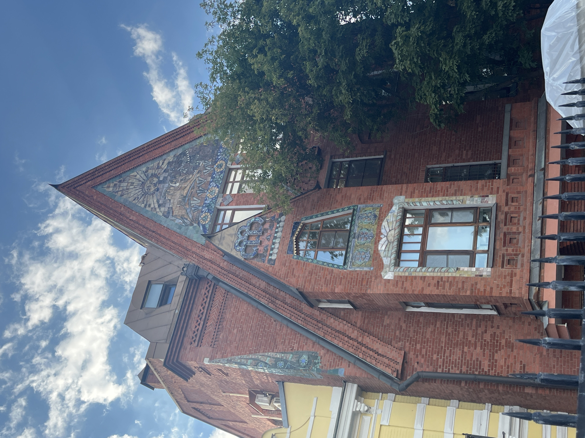 The image shows a multi-story red brick building with decorative mosaic panels on its facade, located in Moscow, Russia. The building has a steeply pitched roof and features ornate windows with intricate frames. A large green tree partially obscures the right side of the building. In the foreground, a black metal fence with pointed finials is visible, and a white tarp covers an object behind the fence. The sky above is blue with scattered white clouds. The building is likely in an older, historic district of Moscow, given its architectural style.
