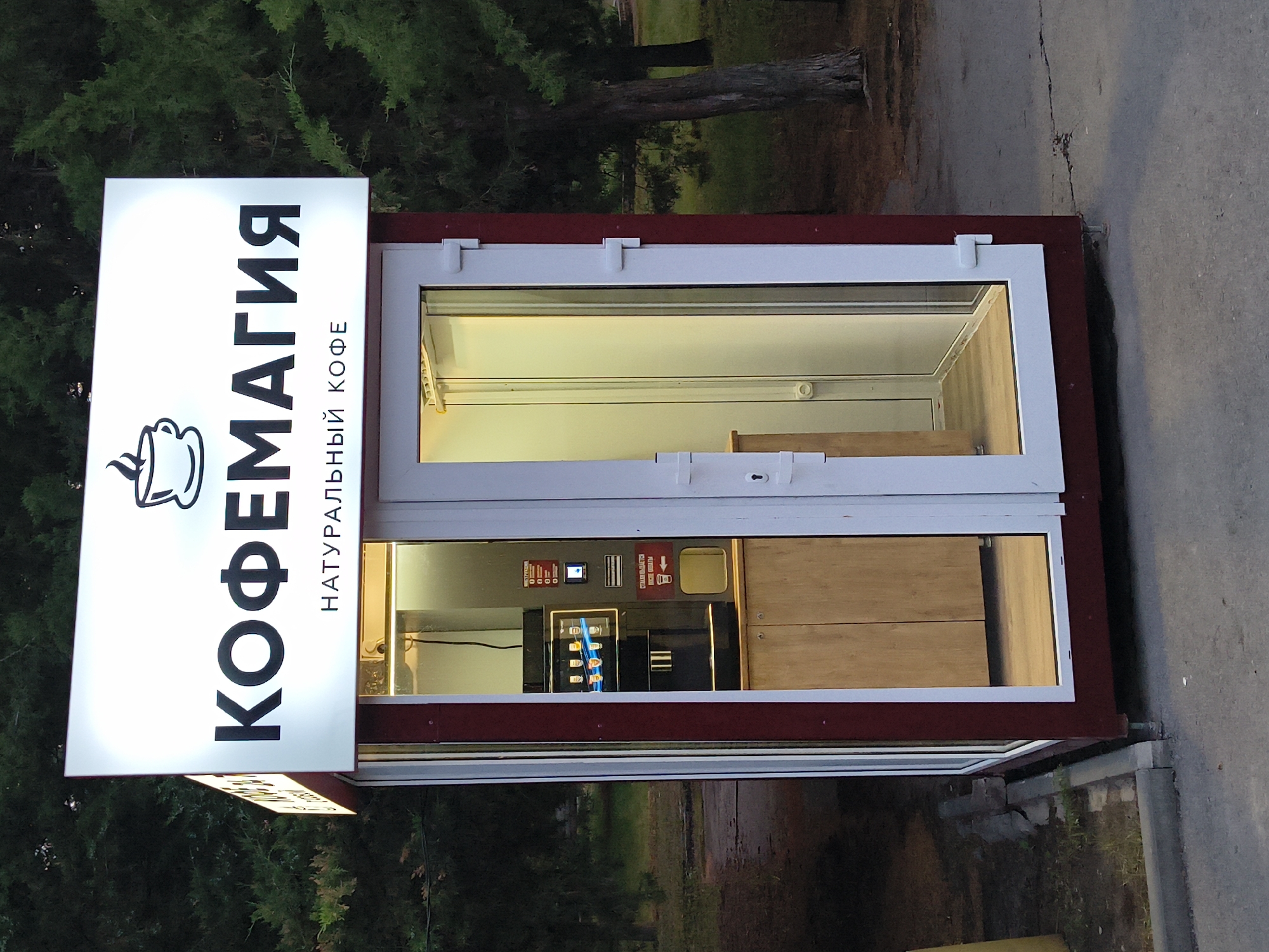 The image shows a coffee kiosk labeled "КОФЕМАГИЯ" (KOFEMAGIA) and "НАТУРАЛЬНЫЙ КОФЕ" (NATURAL COFFEE) in Mikhaylovka, Russia. The kiosk is a small, rectangular structure with a white illuminated sign at the top displaying the name and a coffee cup logo. The kiosk has a dark red frame and glass doors. Inside, a coffee machine is visible, along with some wooden cabinets. The kiosk is situated outdoors, with trees and foliage in the background, suggesting a park or a street with greenery. The ground in front of the kiosk appears to be paved.