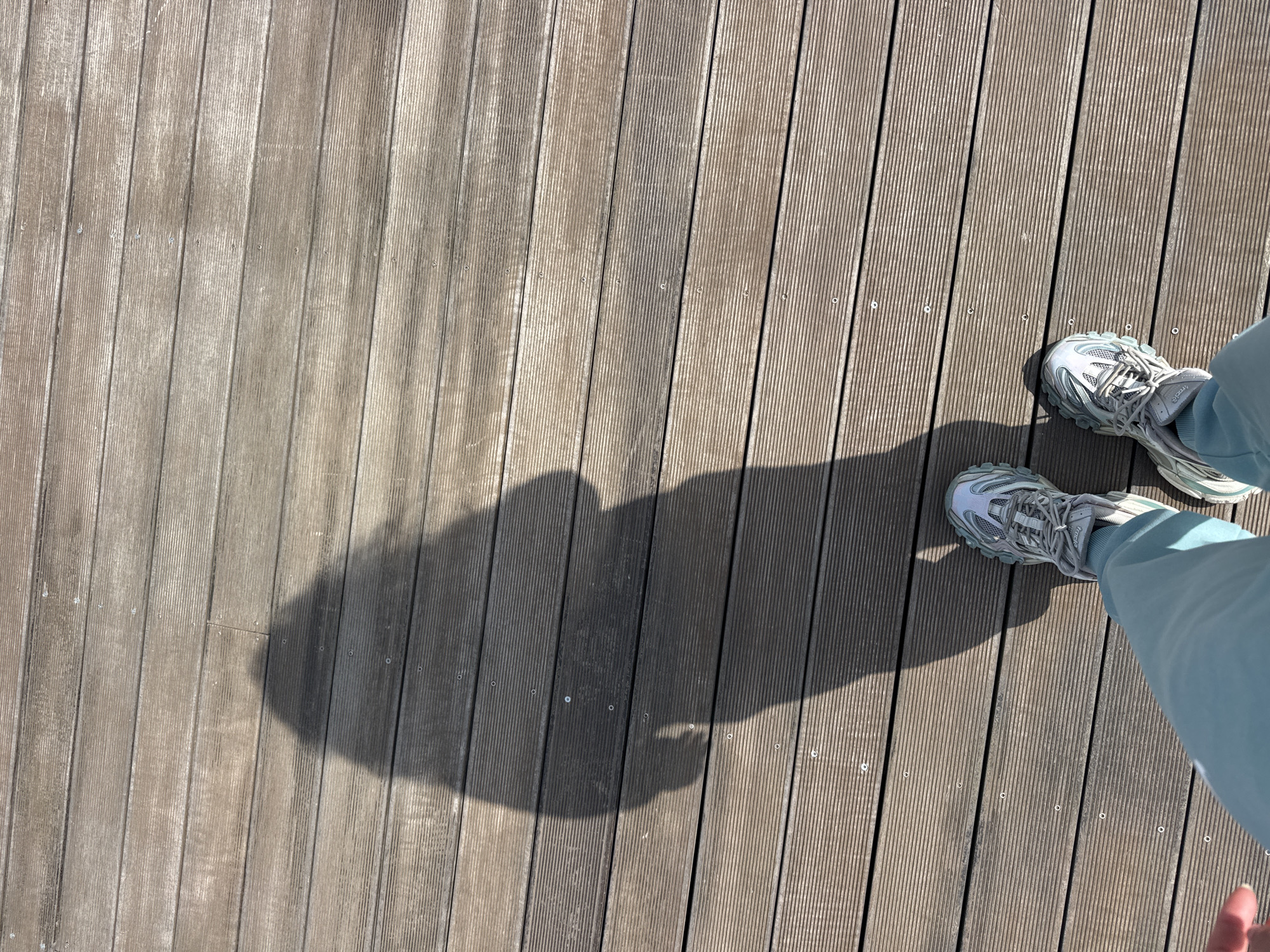 The image shows a pair of feet wearing grey sneakers and light blue trousers standing on a wooden deck. The deck is made of parallel wooden planks, and the shadow of a person is cast on the planks. The scene appears to be outdoors during the daytime. It is impossible to determine the precise location in Sofia, Bulgaria, from the visual information provided.