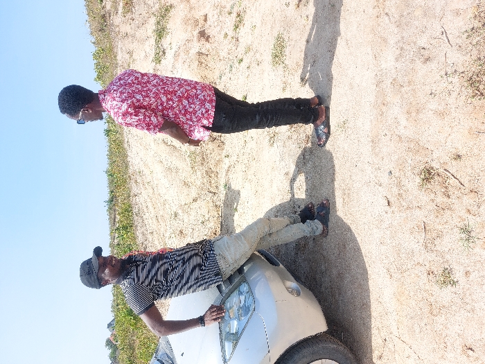 Two adult males are positioned outdoors in a dry, open area within Kibaha, Tanzania. A silver sedan occupies the left foreground, with its front driver's side partially visible. The male on the left, wearing a black baseball cap, a black and white striped short-sleeved shirt, light-colored trousers, and dark sandals, leans against the car's hood/fender, his right hand resting on the surface. He is bearded and smiles, looking towards the second male. The male on the right, wearing dark sunglasses, a red and white patterned short-sleeved shirt, dark trousers, and patterned sandals, stands facing the first male, his hands clasped in front. The ground consists of light brown, sandy dirt with sparse green vegetation. Distinct shadows are cast, indicating bright sunlight from a high angle. The background features a gentle rise in the terrain, covered with scattered green foliage, under a clear blue sky.