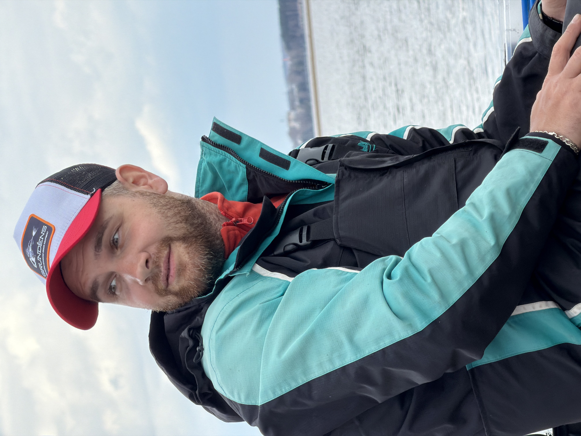 A man with a beard is wearing a baseball cap and a black and turquoise jacket. He is on a boat, and the background shows a body of water and some land. The man is looking towards the camera. The man's hat is a Grundens baseball cap with red brim, and white front panel. He is wearing a silver bracelet on his right wrist. The jacket is a fishing jacket with black panels and turquoise accents. The water behind the man appears to be a lake or river, and there are trees and buildings visible on the far shore. Based on the location, it is possible that the body of water is the Kama River or one of its reservoirs.