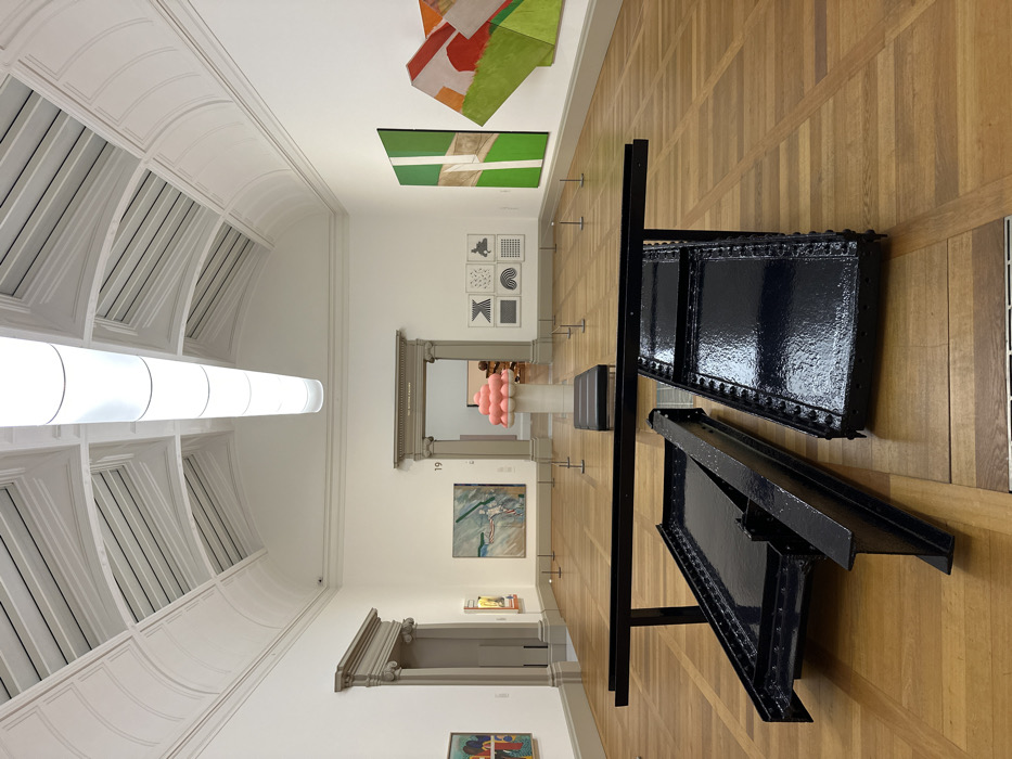 The image displays the interior of an art gallery, specifically identified by signage as Room 19 of The Serpentine Galleries, located in Kensington Gardens, London, United Kingdom. The room is empty of people.
The scene features a light brown parquet floor and white walls. The high, vaulted ceiling is white with exposed structural elements and a long, cylindrical light fixture suspended centrally.
In the foreground, two large, dark blue-black industrial-style metal objects are positioned directly on the floor. One is a flat, rectangular form with visible rivets, and the other is an I-beam structure, with a horizontal bar extending from it. They appear to be sculptural installations.
In the midground, a black padded bench is centered on the floor. Behind it, a white cylindrical pedestal supports a sculpture composed of numerous pink spherical objects clustered into a rounded, irregular form. Along the walls, various contemporary artworks are displayed: on the left, a blue and green abstract painting; on the right, a large vertical green abstract painting with light geometric divisions, and an adjacent multi-panel abstract piece in orange, red, and green. To the left of the pink sculpture, a grid of six smaller framed artworks exhibits black and white geometric patterns.
Two large archways with classical pilasters and cornices are visible. The central archway is open, revealing a continuation of the gallery space, and is labeled "THE SERPENTINE GALLERIES" with the number "19" above it. Small, dark, low-profile stanchions are spaced throughout the midground. The room is brightly illuminated.