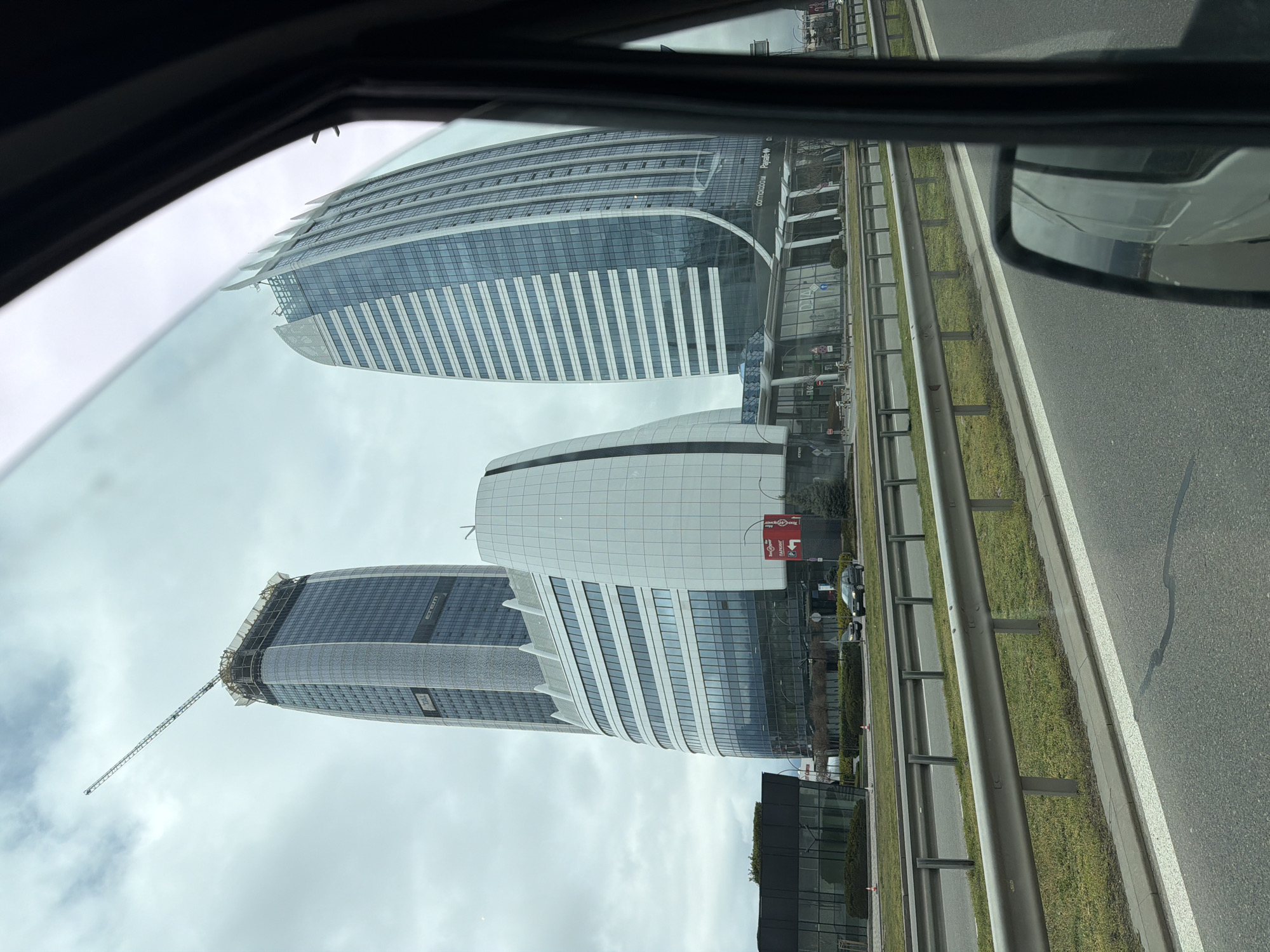 The image shows a view from inside a vehicle, likely a car, looking out towards several modern buildings on a roadside. These buildings are located at "Sofia Airport Center", 1 Christopher Columbus Blvd, Sofia, Bulgaria. The buildings are characterized by their contemporary architecture, featuring curved facades and reflective glass panels. There's a construction crane atop one of the buildings, indicating ongoing development. A grey metal guardrail runs alongside the road. The sky is overcast, and the view is partly obstructed by the vehicle's frame and side mirror.