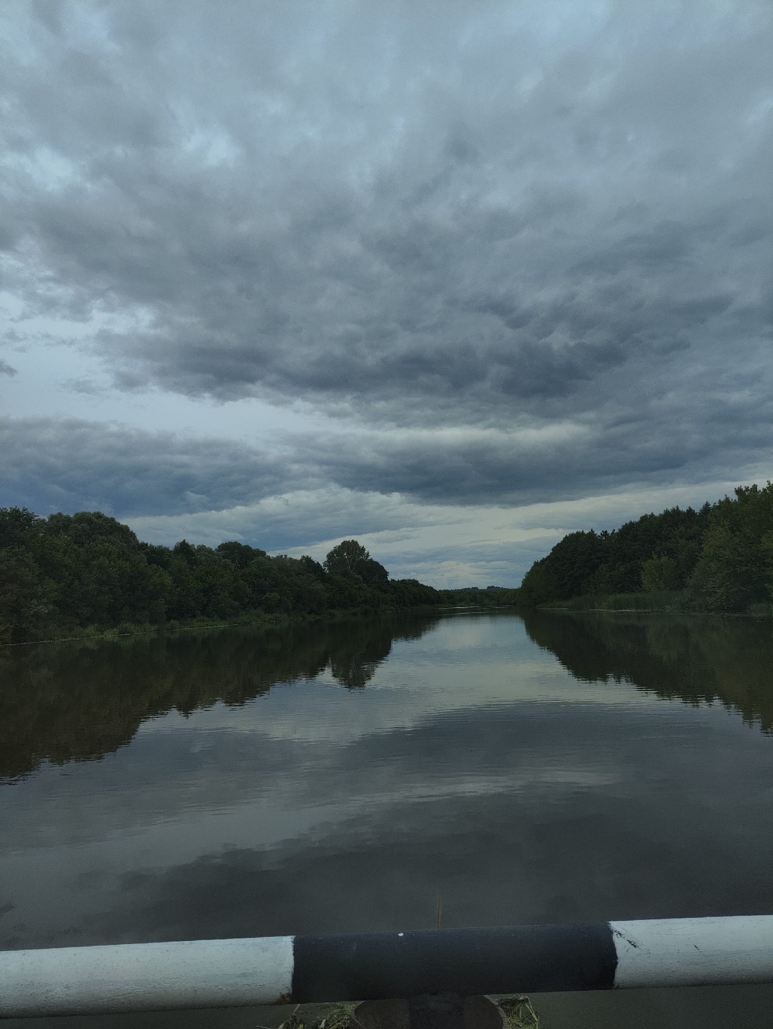 This is a full-frame, eye-level, medium shot of a wide, calm river. The river dominates the lower half of the frame, reflecting the dark, cloudy sky and the dense green treeline that borders its banks. The sky is filled with heavy, gray storm clouds, creating a somber atmosphere. The treeline stretches across the horizon, with lush foliage visible on both sides of the river. The foreground of the image features the top of a metal railing, painted white and black, which is partially obscuring the lower edge of the scene. The overall impression is one of natural beauty under an impending storm. The location is identified as the river in Tumutuk, Russia.