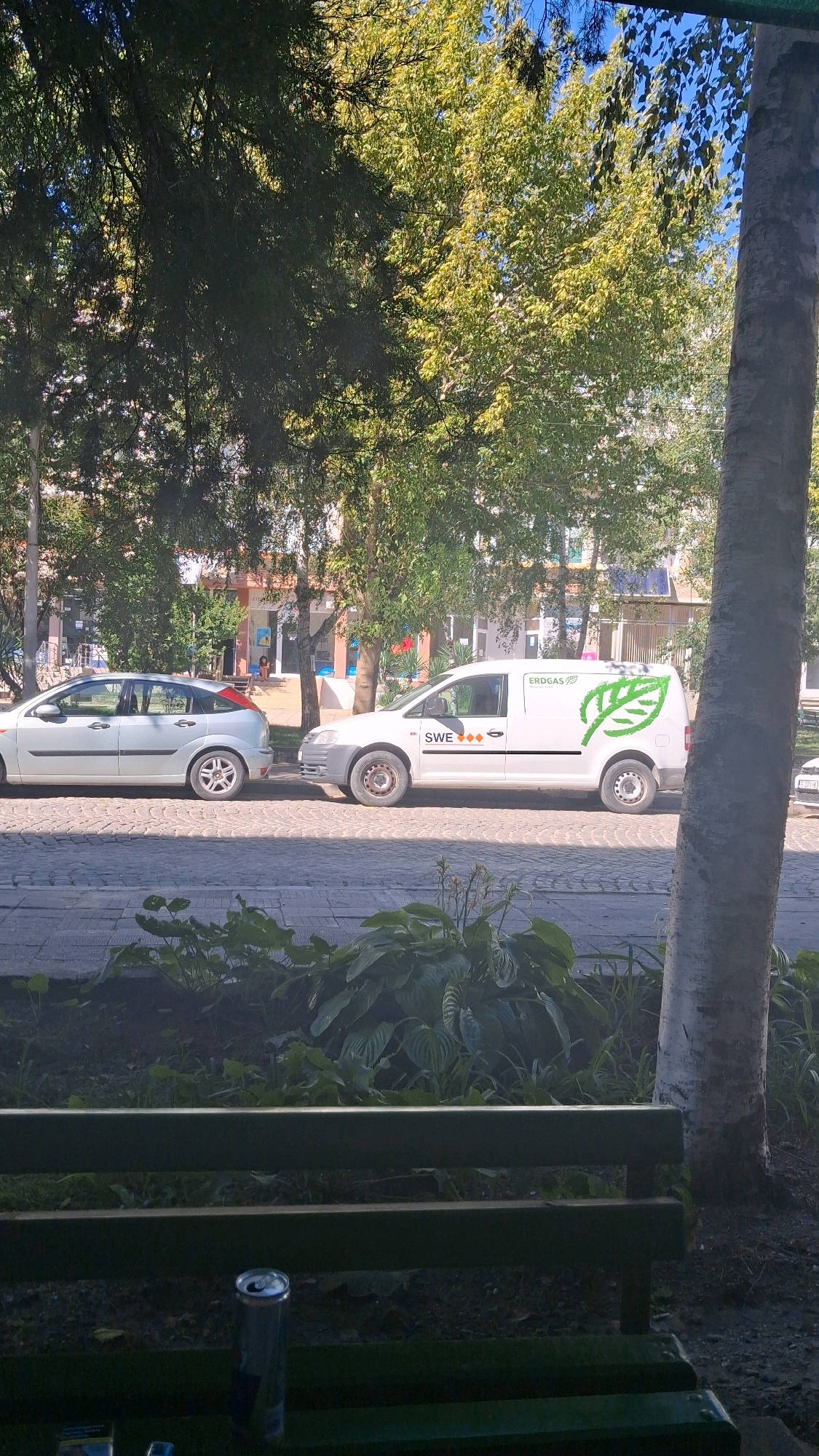 In Gotse Delchev, Bulgaria, on a sunny day, a light-colored hatchback car is parked on a cobblestone street, facing left. Adjacent to it, a white utility van with the "ERDGAS" logo and a green leaf emblem is also parked, facing right. The van has "SWE" and three orange diamonds on its side. Behind the vehicles, trees with green foliage line the street, with buildings visible in the background. In the foreground, a dark green park bench sits in front of dense green plants and a tree trunk on the right. A silver aluminum can is visible on the bench.