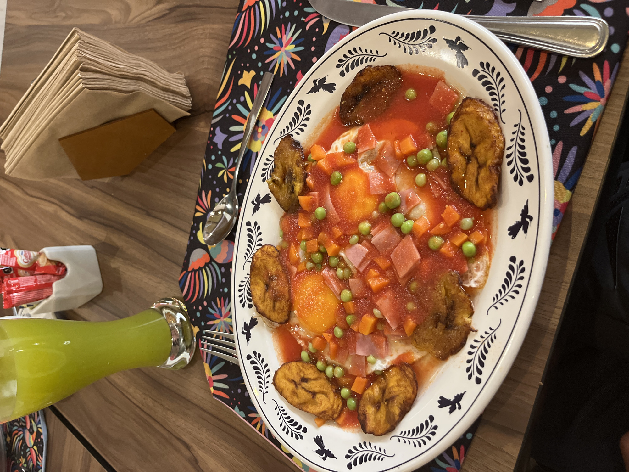 On a wooden table, a plate of food sits on a colorful patterned placemat. The plate is white with black leaf and bird designs around the rim. The dish features fried eggs, plantains, diced ham, peas, and diced carrots in a red sauce. To the left, there is a stack of brown napkins, a white container with red condiments, and a green drink in a glass bottle. Silverware is visible near the plate on the placemat. The image's location is Unknown, Unknown.