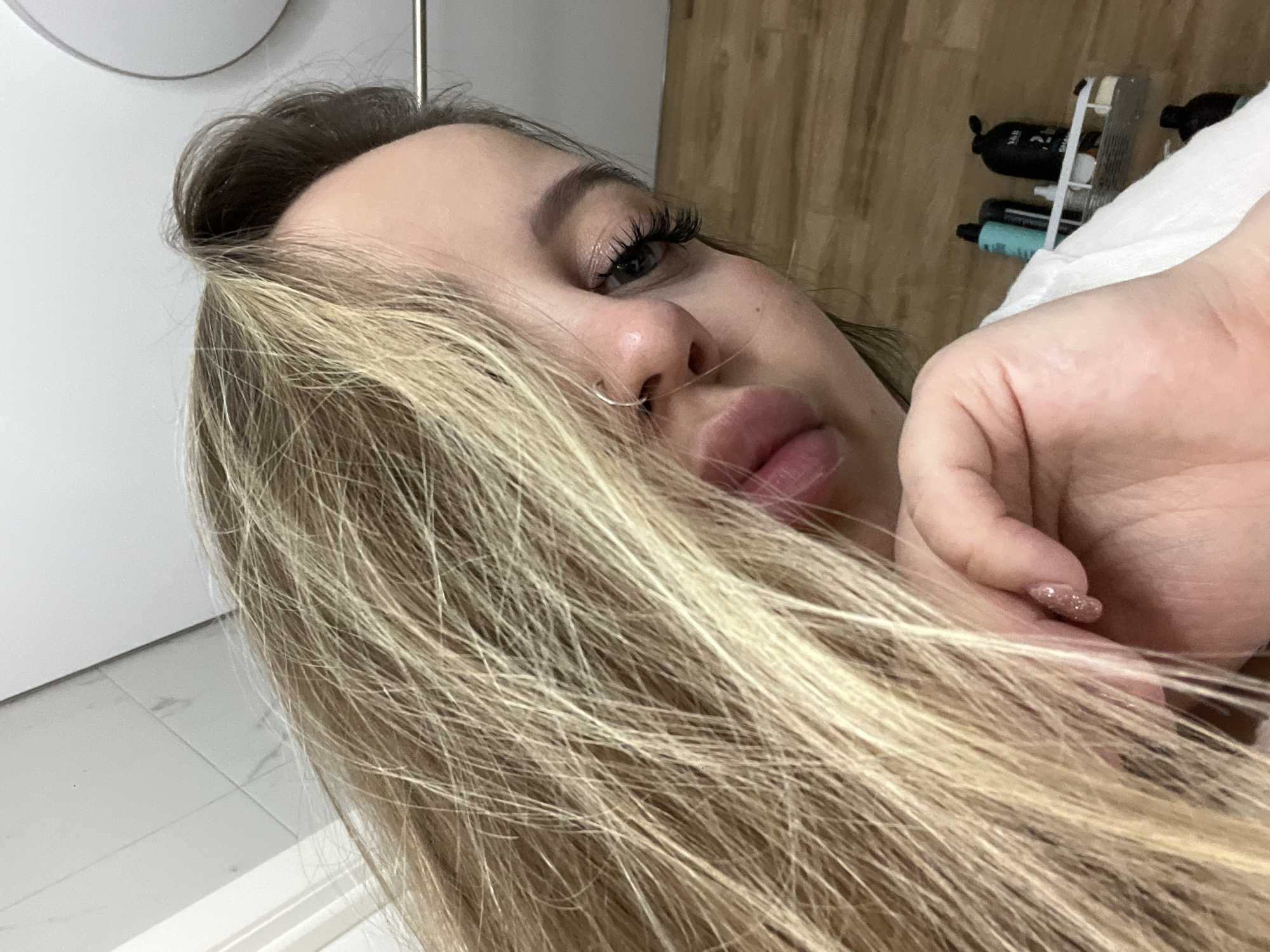 A close-up, angled portrait features a person, likely female, lying down indoors in Krasnodar, Russia. The subject's face is partially obscured by long, flowing light blonde hair, with darker roots visible near the scalp. Her left eye is visible, characterized by prominent long eyelashes and well-defined eyebrows. A silver hoop nose ring adorns her left nostril, and her full lips are slightly parted.
Her right hand is positioned near her face and hair, revealing fingernails painted with a glittery pink or gold polish. The background is partially blurred but provides contextual cues: to the left, there is a plain white wall and a floor covered with white tiles. To the right, behind the person's hand, several personal care product bottles are arranged on a shelf, including a black bottle and a teal-colored one. A section of wooden wall paneling is also visible, and a white fabric, possibly a towel or bedding, can be seen. The image captures a moment of relaxation. No readable text is visible on any objects.