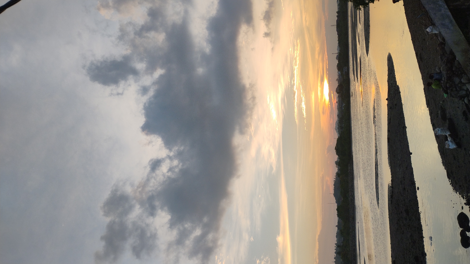 This is a vertical, eye-level, medium shot of a sunset over a river in Lhokseumawe, Indonesia. The sky is filled with dramatic clouds, ranging from light grey to dark grey, with patches of soft orange and yellow hues from the setting sun. The sun itself is a bright, concentrated orb just above the horizon line.
The foreground is dominated by a muddy riverbank, with patches of dark, wet soil and some scattered rocks. There are two figures in the lower right corner of the image, both seated. One appears to be wearing a green shirt, and the other a dark shirt. Several white sacks are also visible on the riverbank.
The river itself is calm, reflecting the colors of the sky. Its surface is broken by several sandbars or exposed mudflats. In the middle ground, a line of trees forms a dark silhouette against the brighter sky. Beyond the trees, the distant landscape is hilly and appears to be populated with some structures and power lines. The overall impression is one of a peaceful, natural scene during twilight.